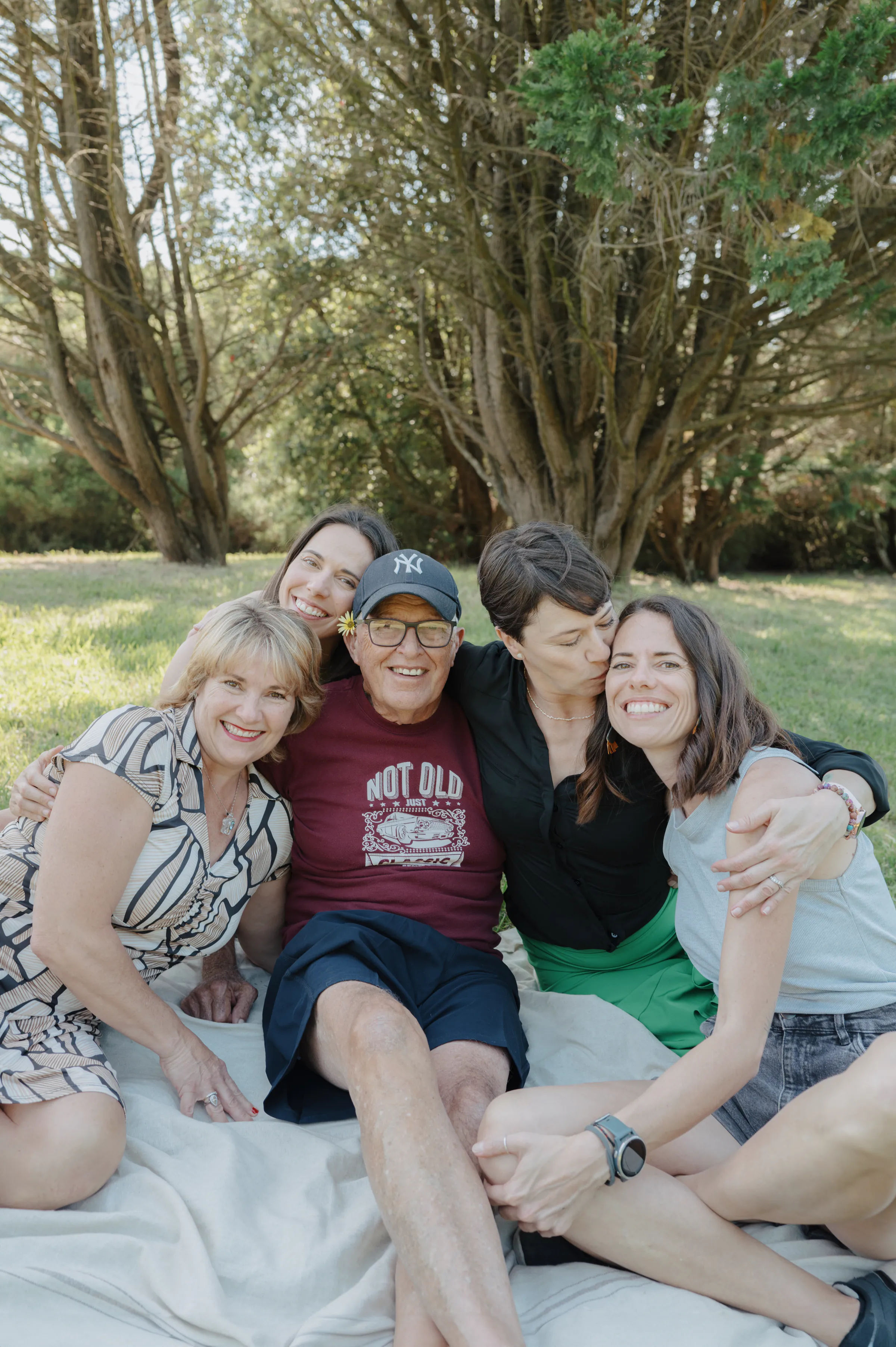 Group portrait of four adults sitting close on a blanket during a soft light park session.