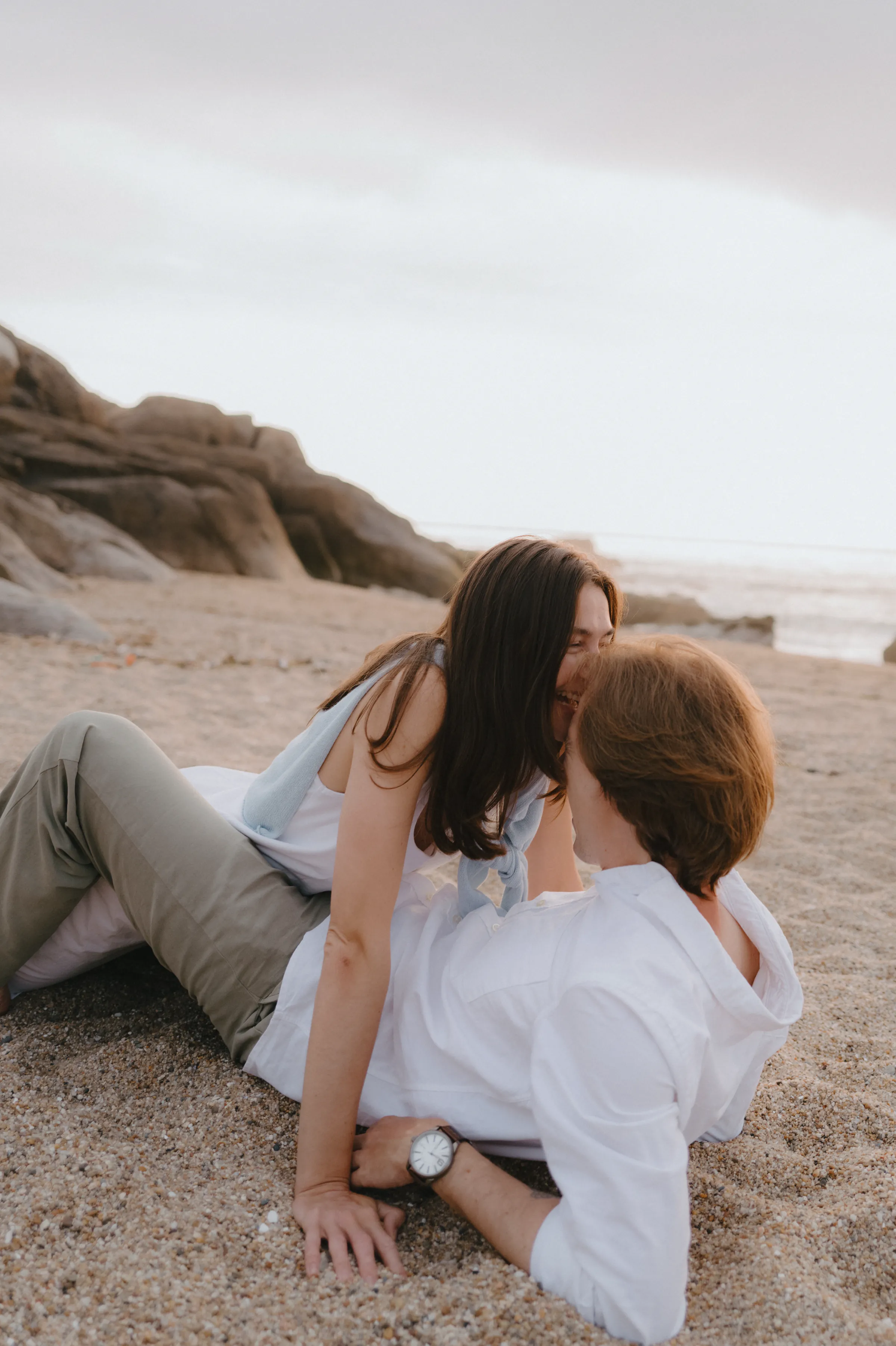 Couple sitting closely on a sandy seaside during an outdoor session in soft overcast light.