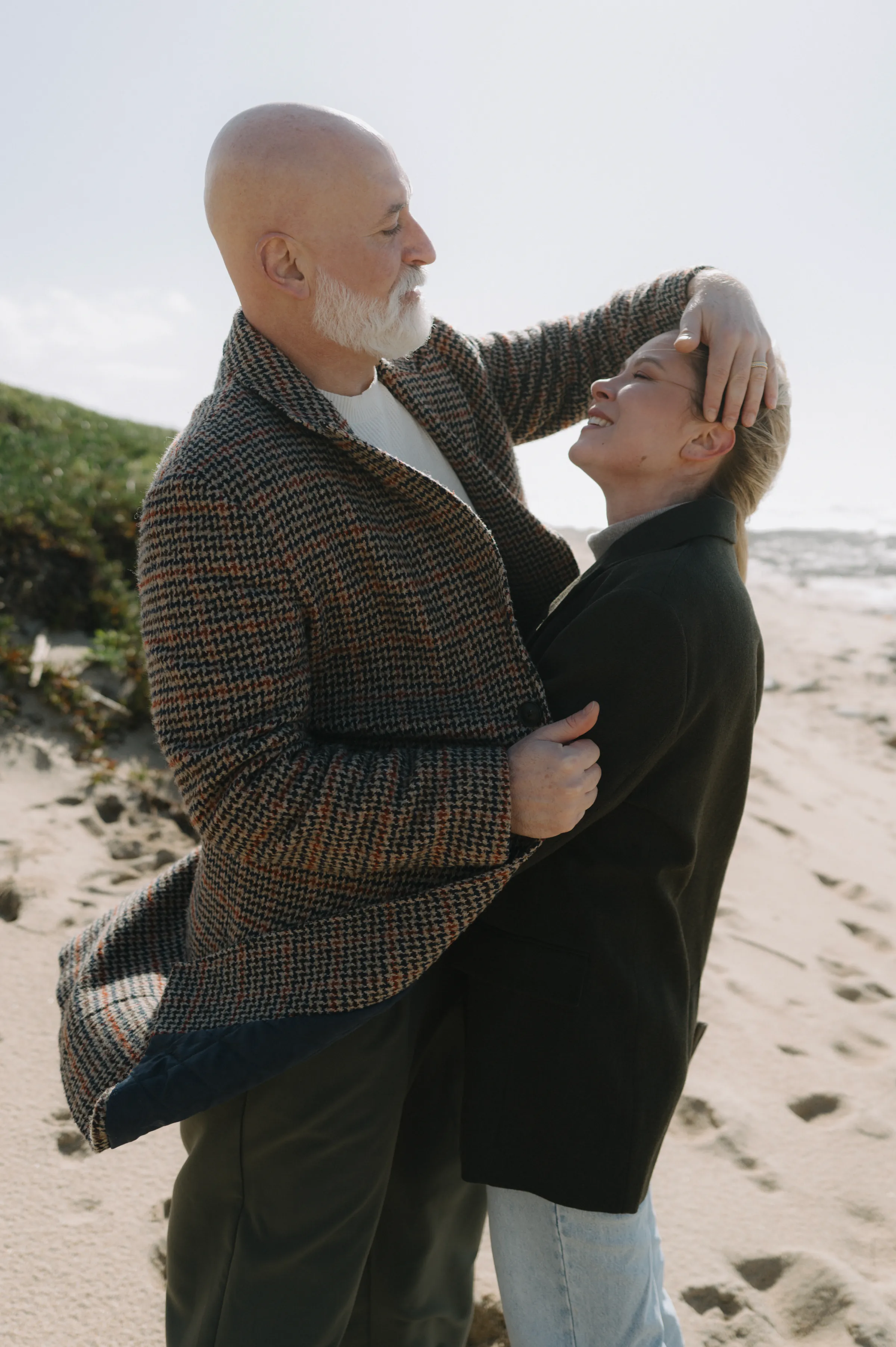 Couple standing close on a sandy beach during a seaside outdoor session in soft light.