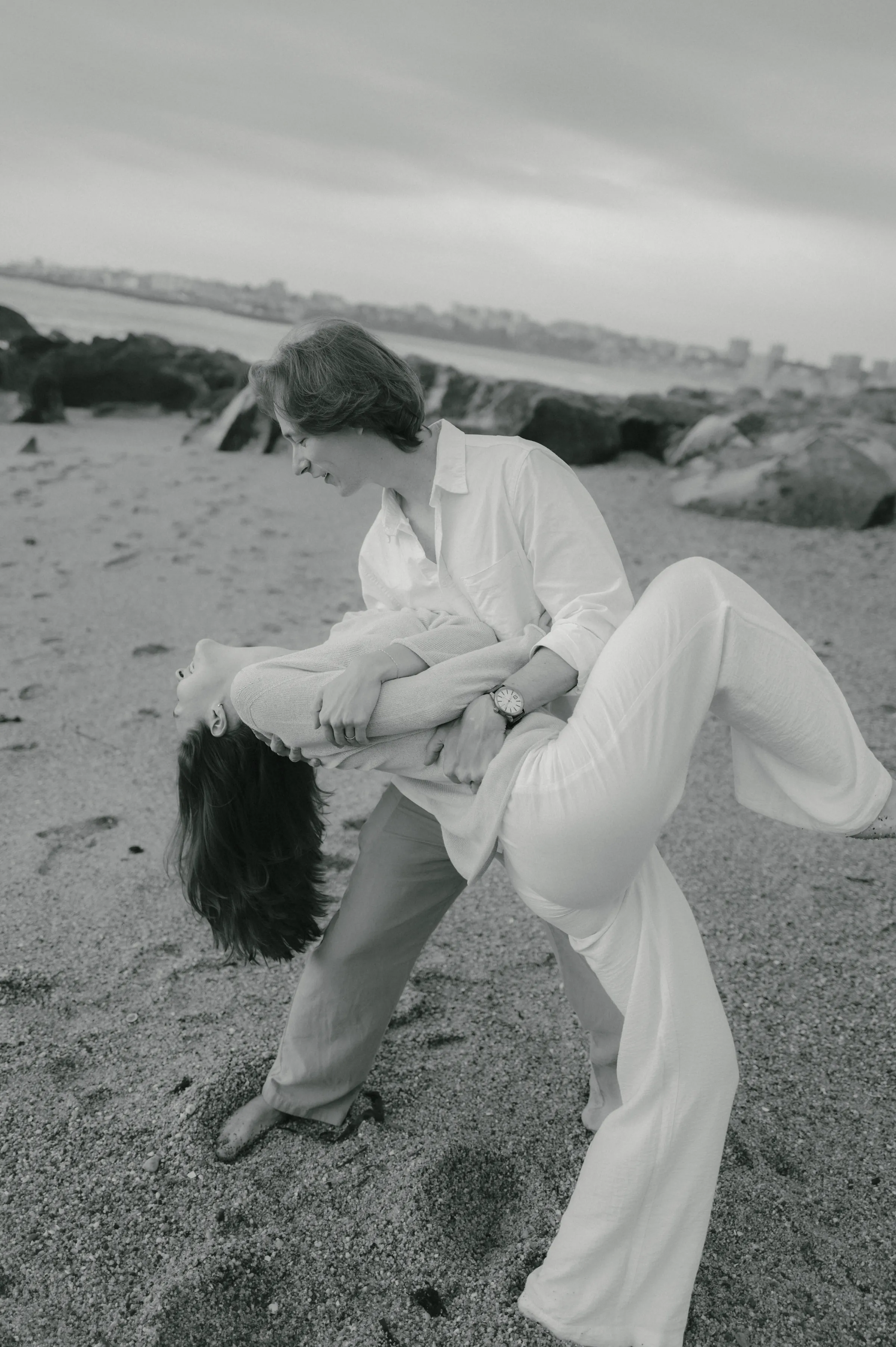 Black-and-white seaside portrait of a couple posing on a sandy beach with rocky coastline behind them.