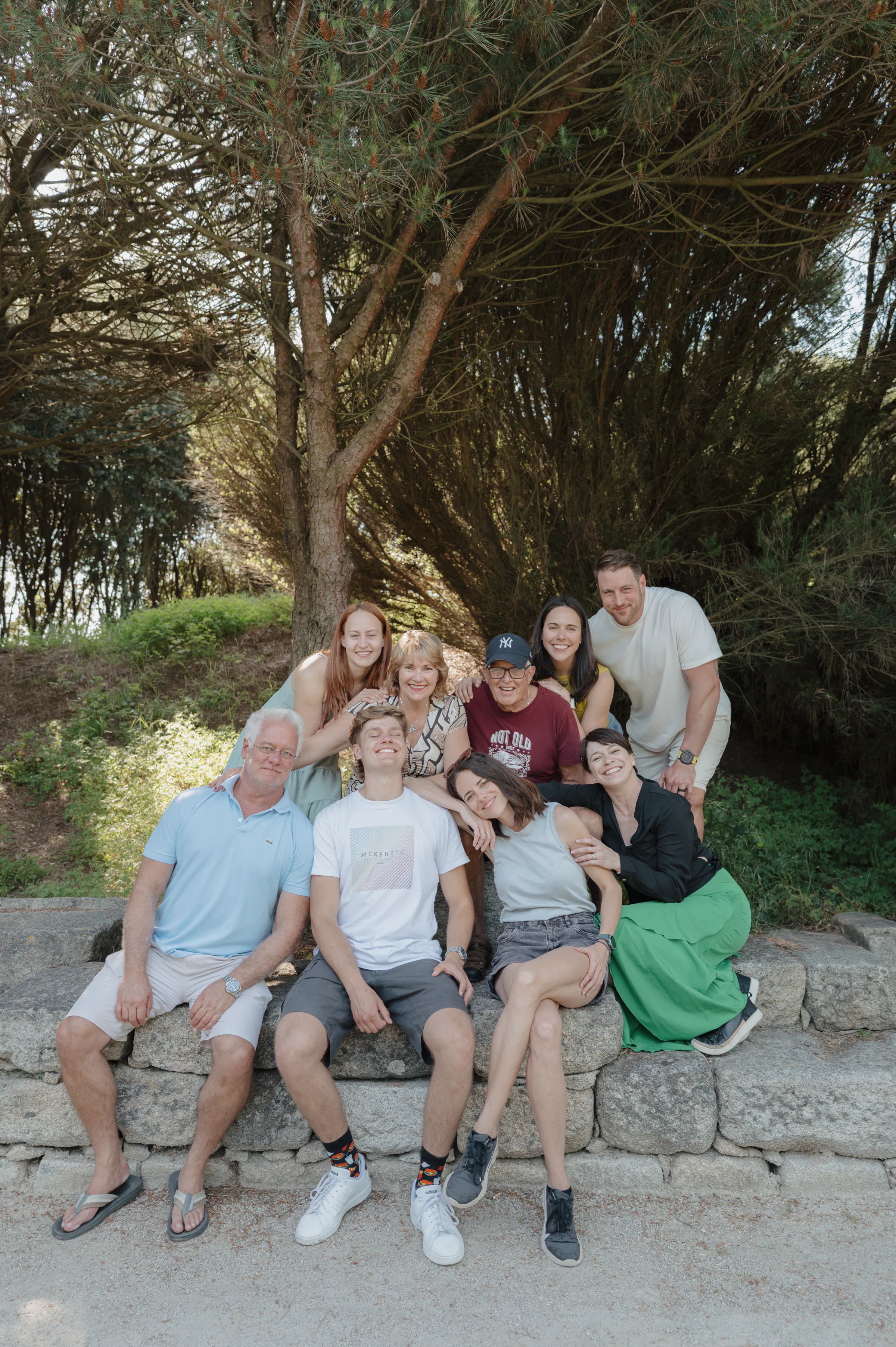 Multi-generational group portrait on a stone wall beneath trees during an outdoor session.