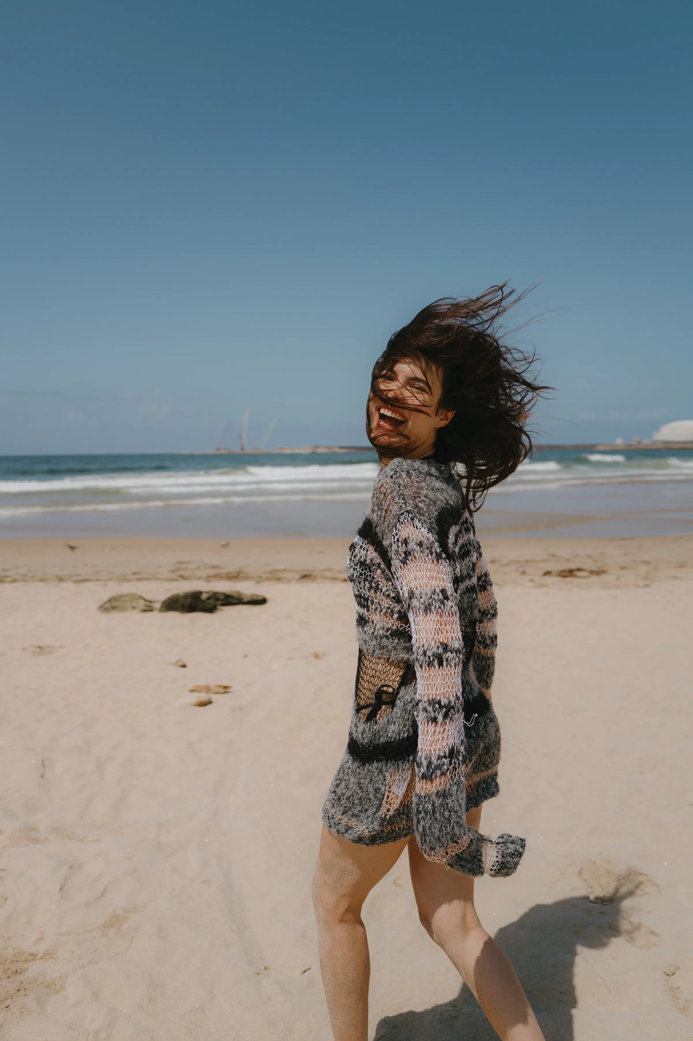 Seaside portrait of a person in a patterned sweater, hair blowing in the wind on a sandy beach.