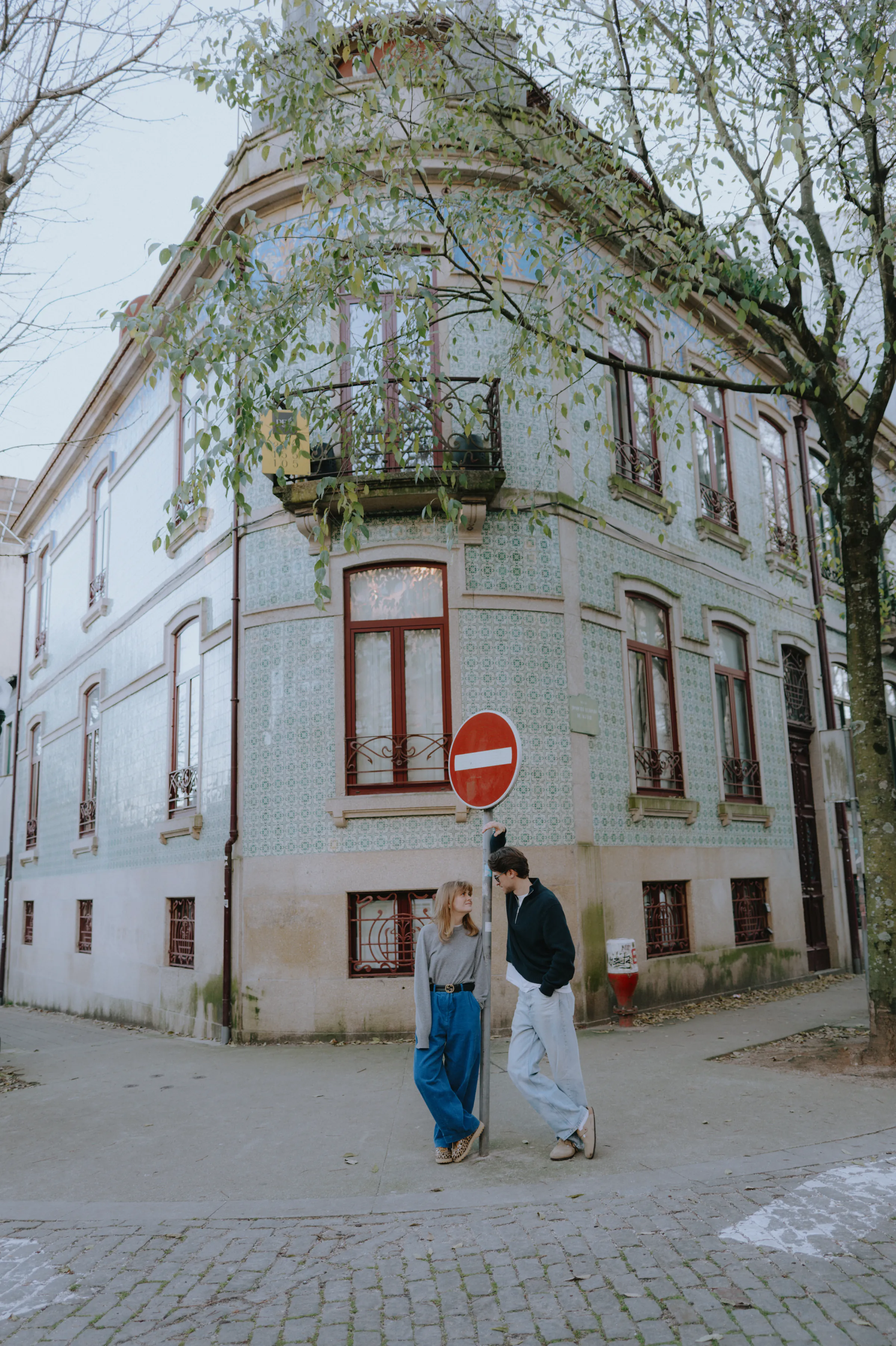 Couple during a city walk beside a tiled corner building and tree-lined street in soft daylight.