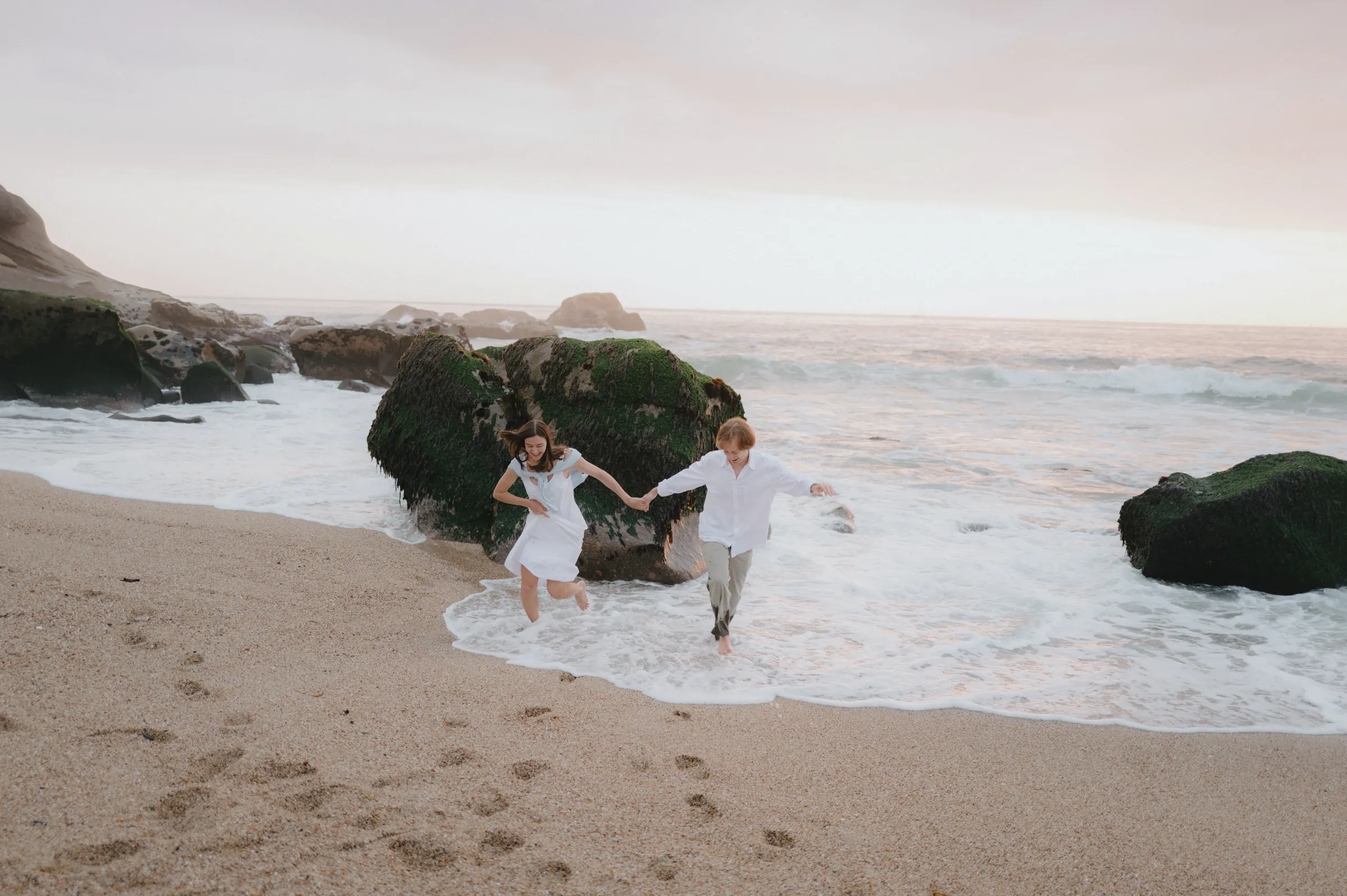 Couple holding hands during a seaside outdoor session, running along the shore near dark rocks and surf.