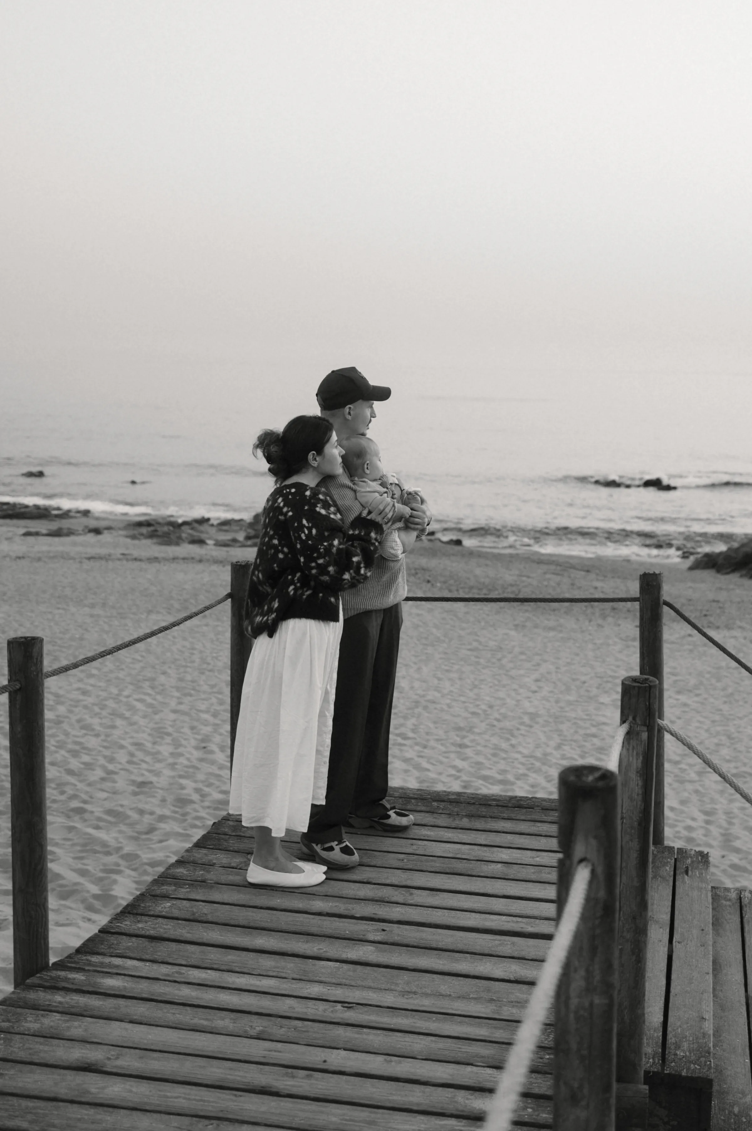Couple standing close on a wooden seaside boardwalk, looking out toward calm water in soft light.