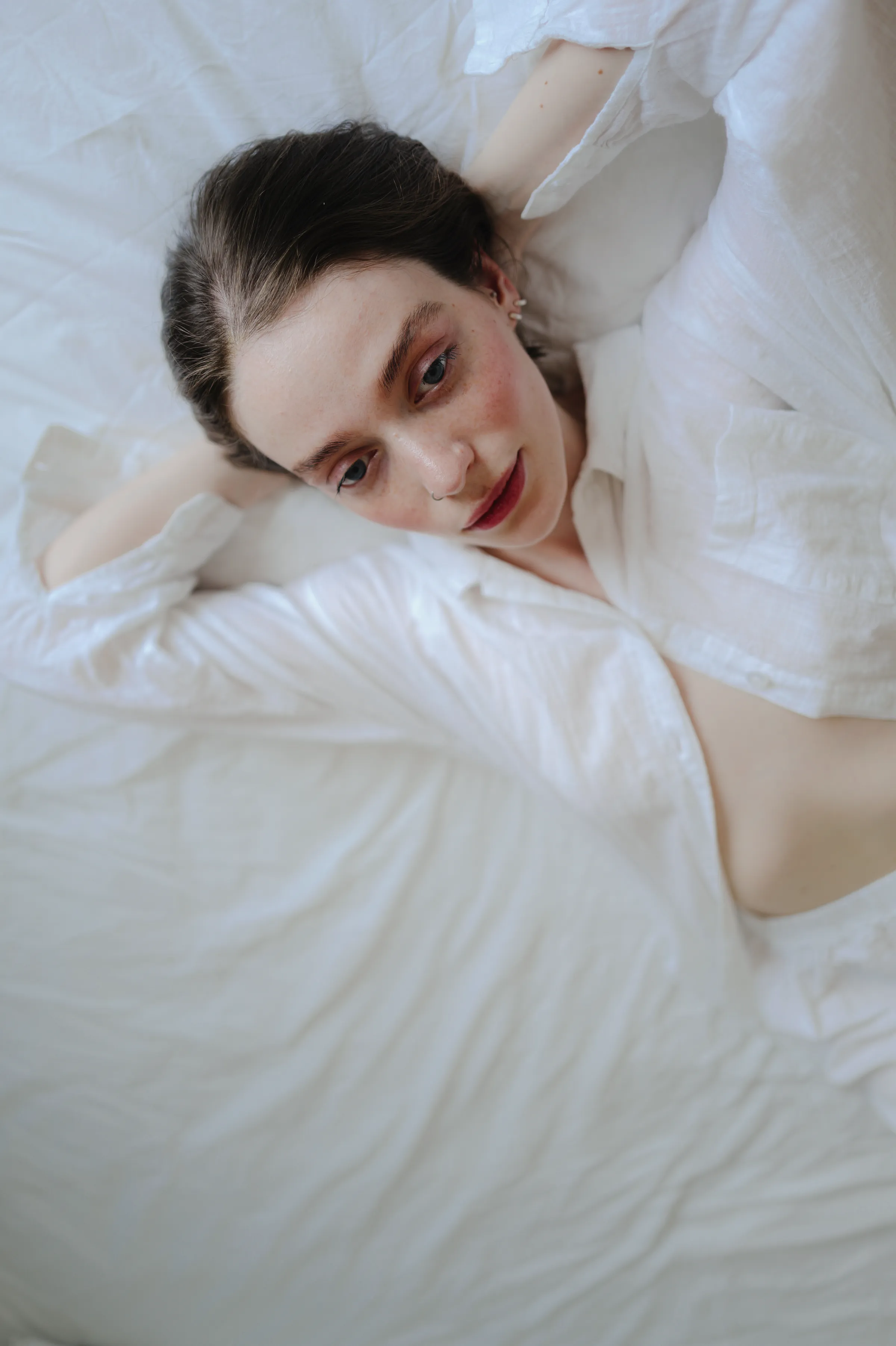 Close portrait of a person reclining on white bedding in soft indoor light.