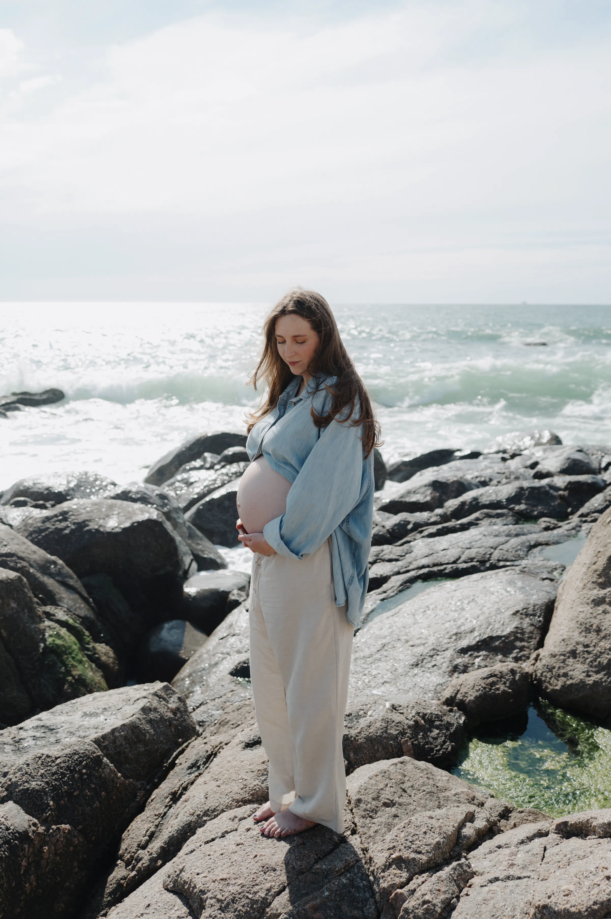 Portrait of a person standing barefoot on seaside rocks with waves behind in soft light.
