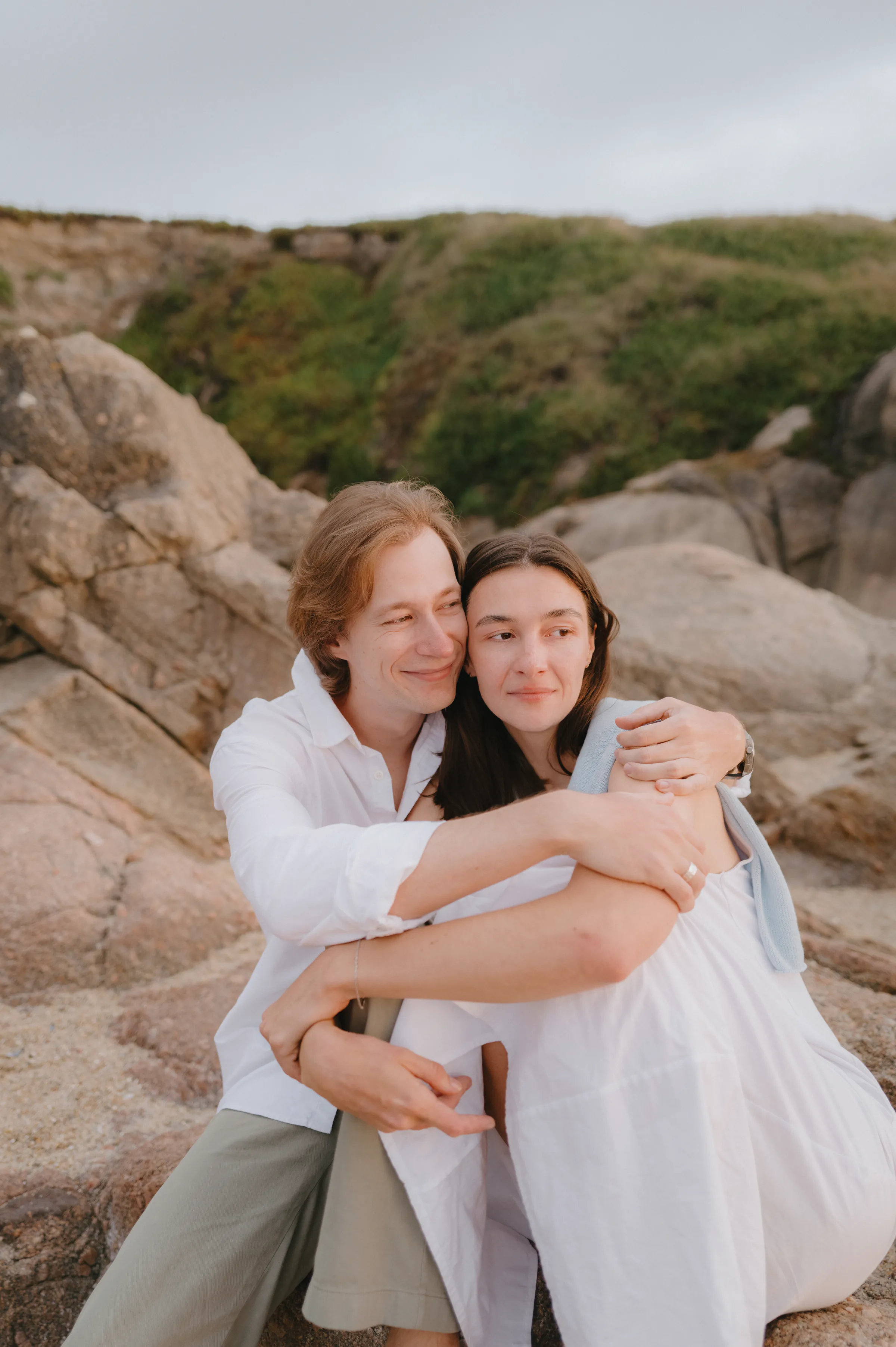 Couple embracing during an outdoor portrait session on rocky coastal terrain with soft overcast light.