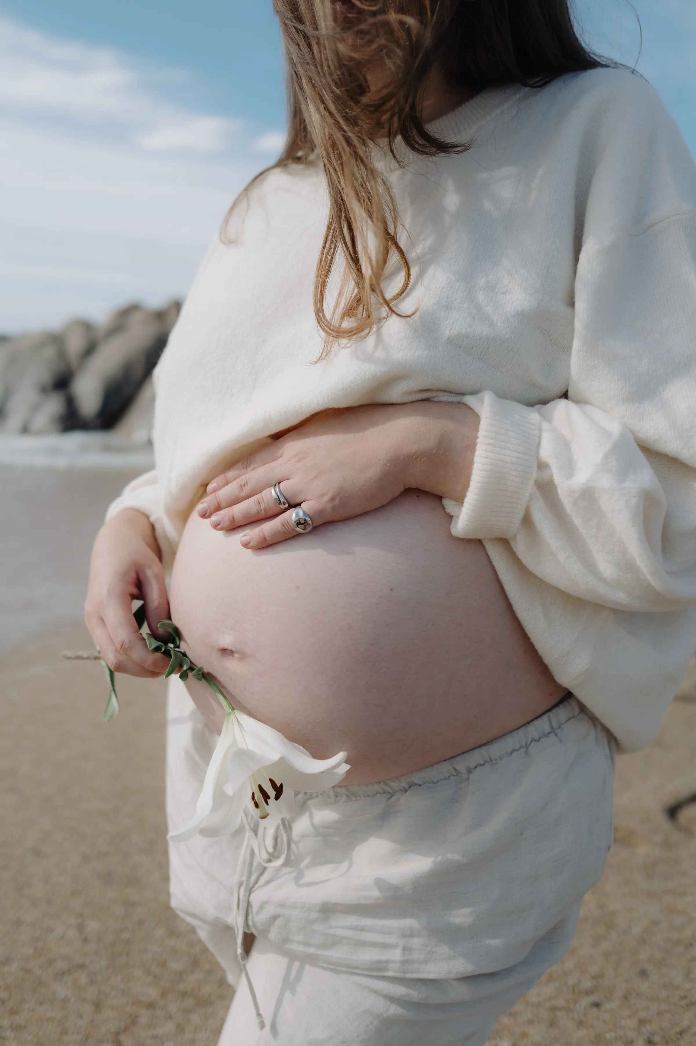 Person in a white sweater holding a bare belly during a seaside outdoor session.