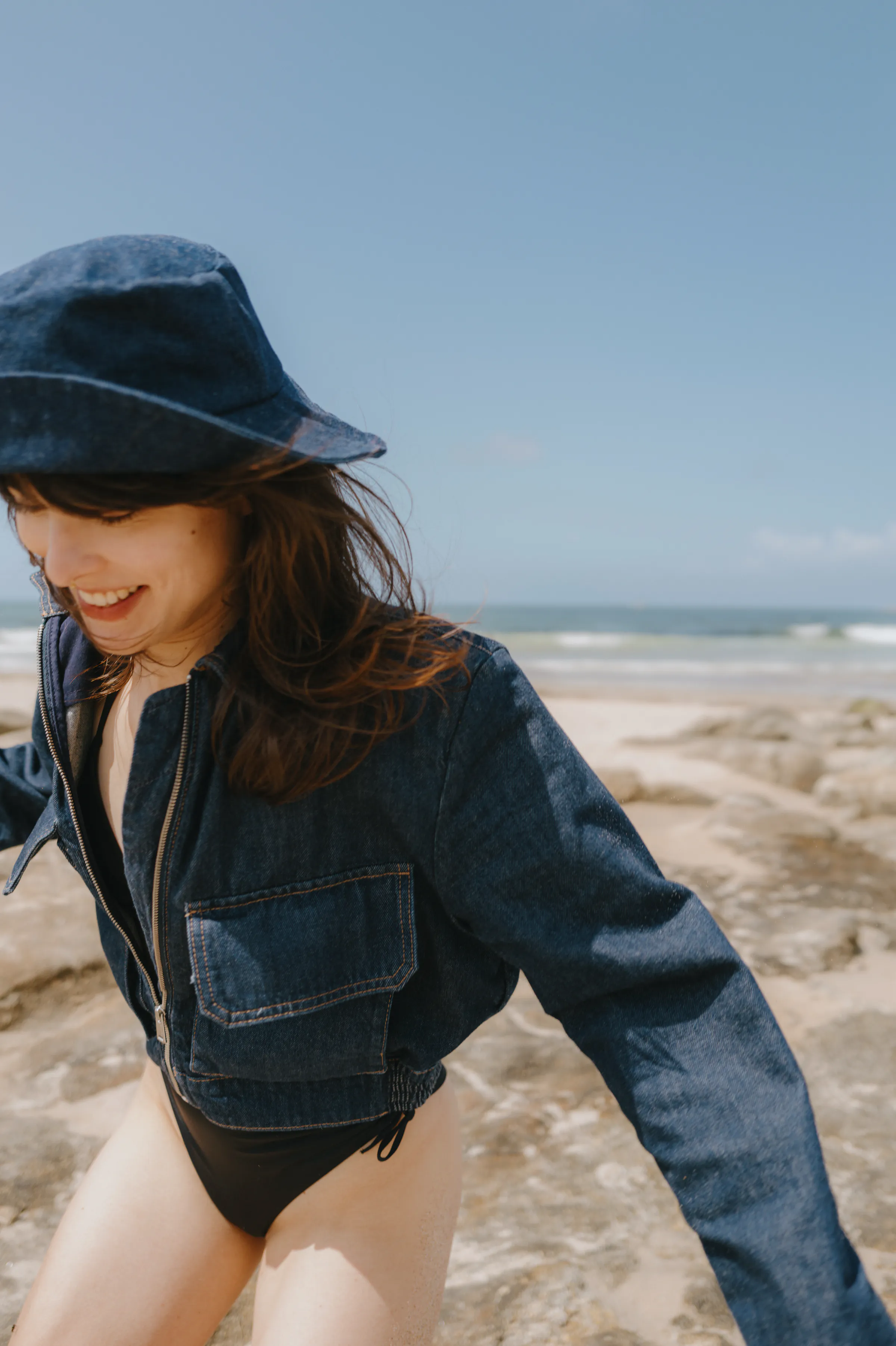 Seaside portrait of a woman in a denim hat and cropped jacket, with ocean waves behind her