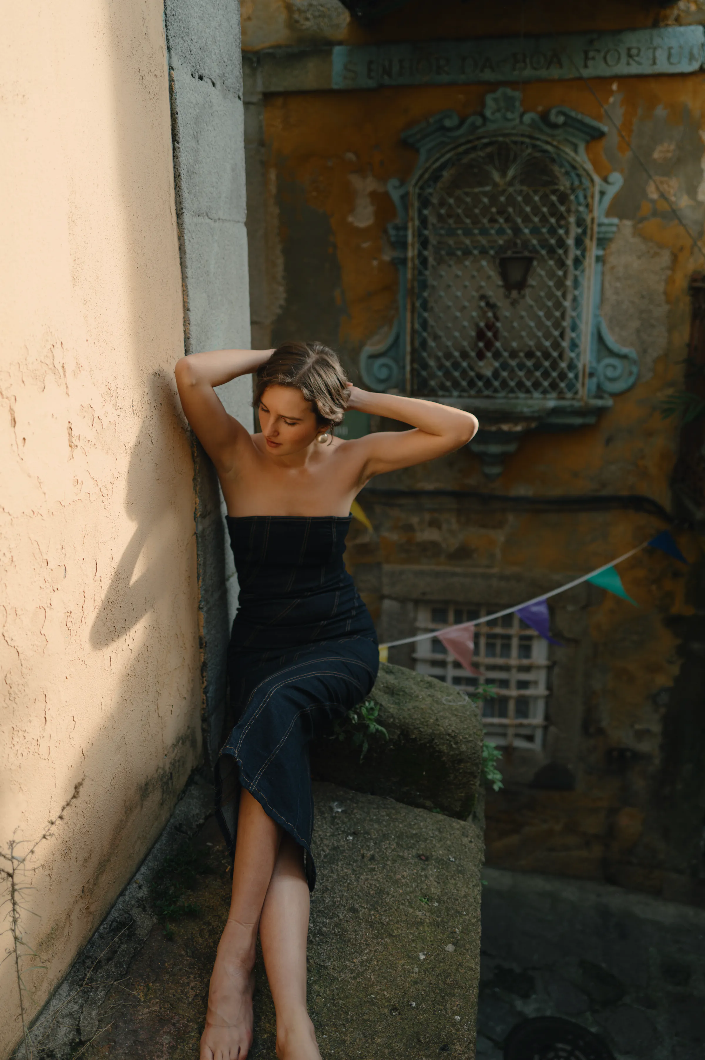 Outdoor portrait of an adult seated on a stone ledge beside a sunlit old wall.