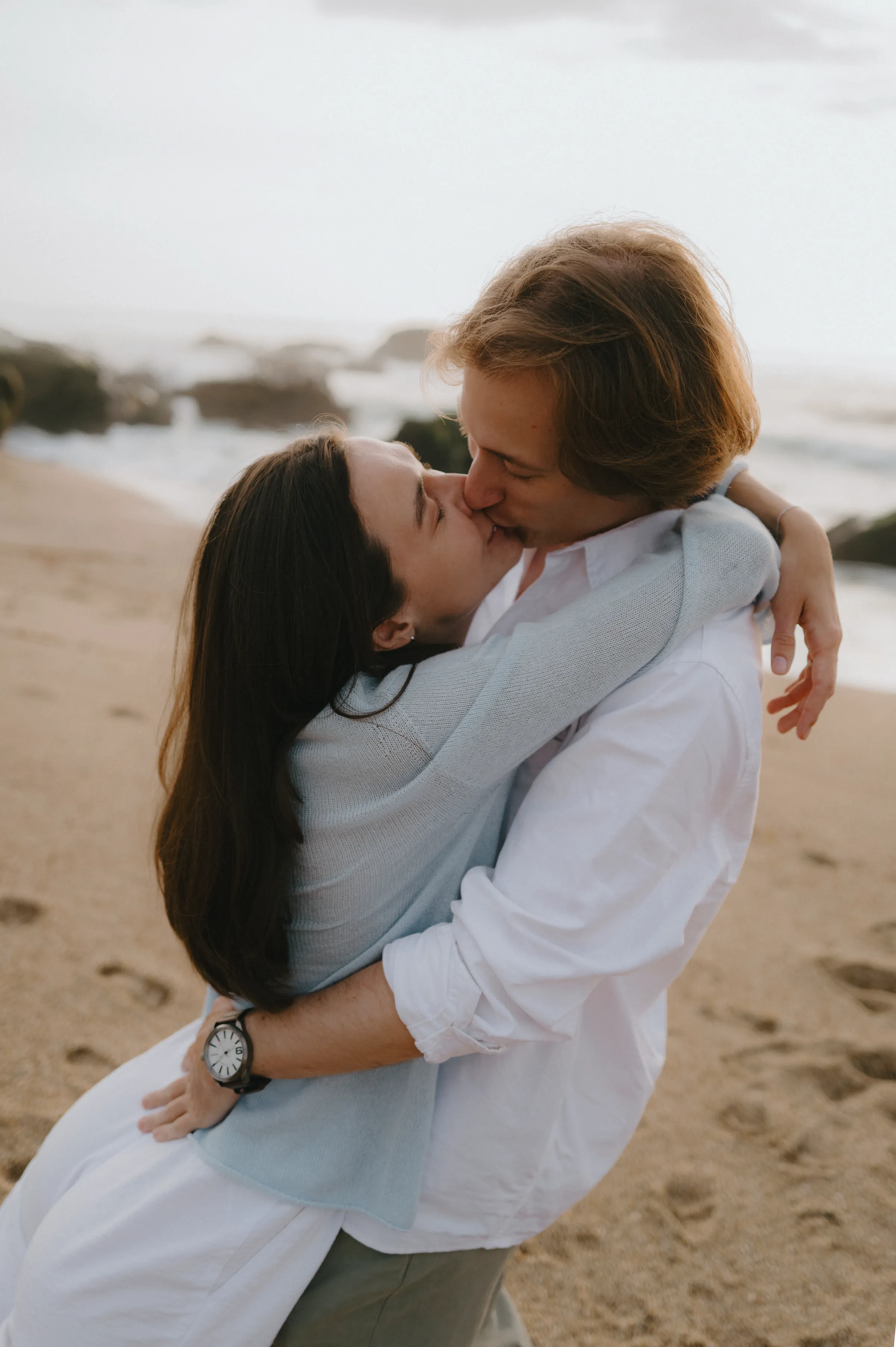 Couple embracing and kissing during a soft light seaside session on a sandy beach.