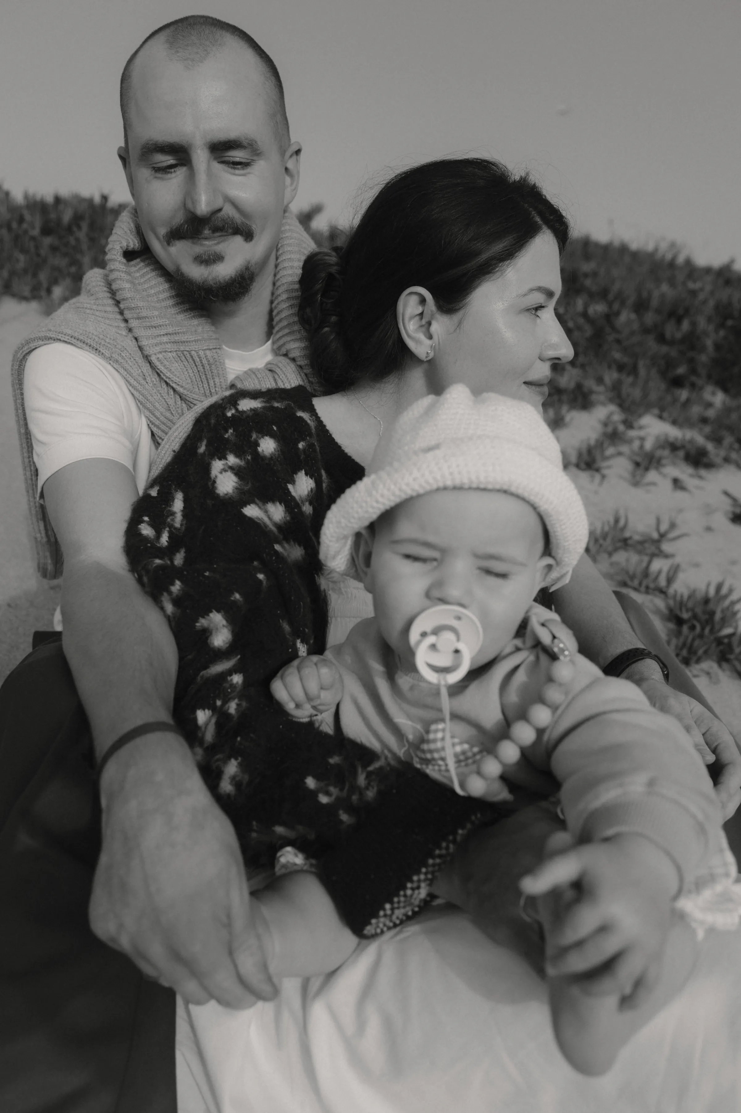 Black-and-white family portrait during an outdoor session, with a baby in a knit hat held close.