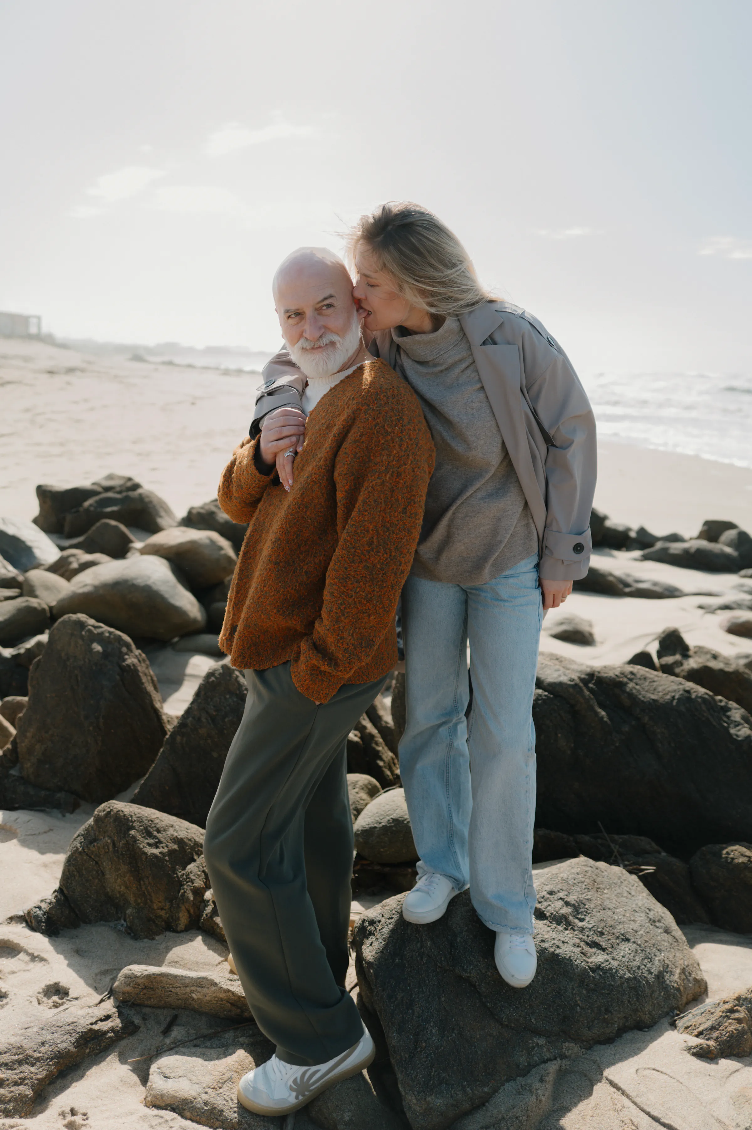 Couple portrait on seaside rocks, standing close together in soft light by the beach.