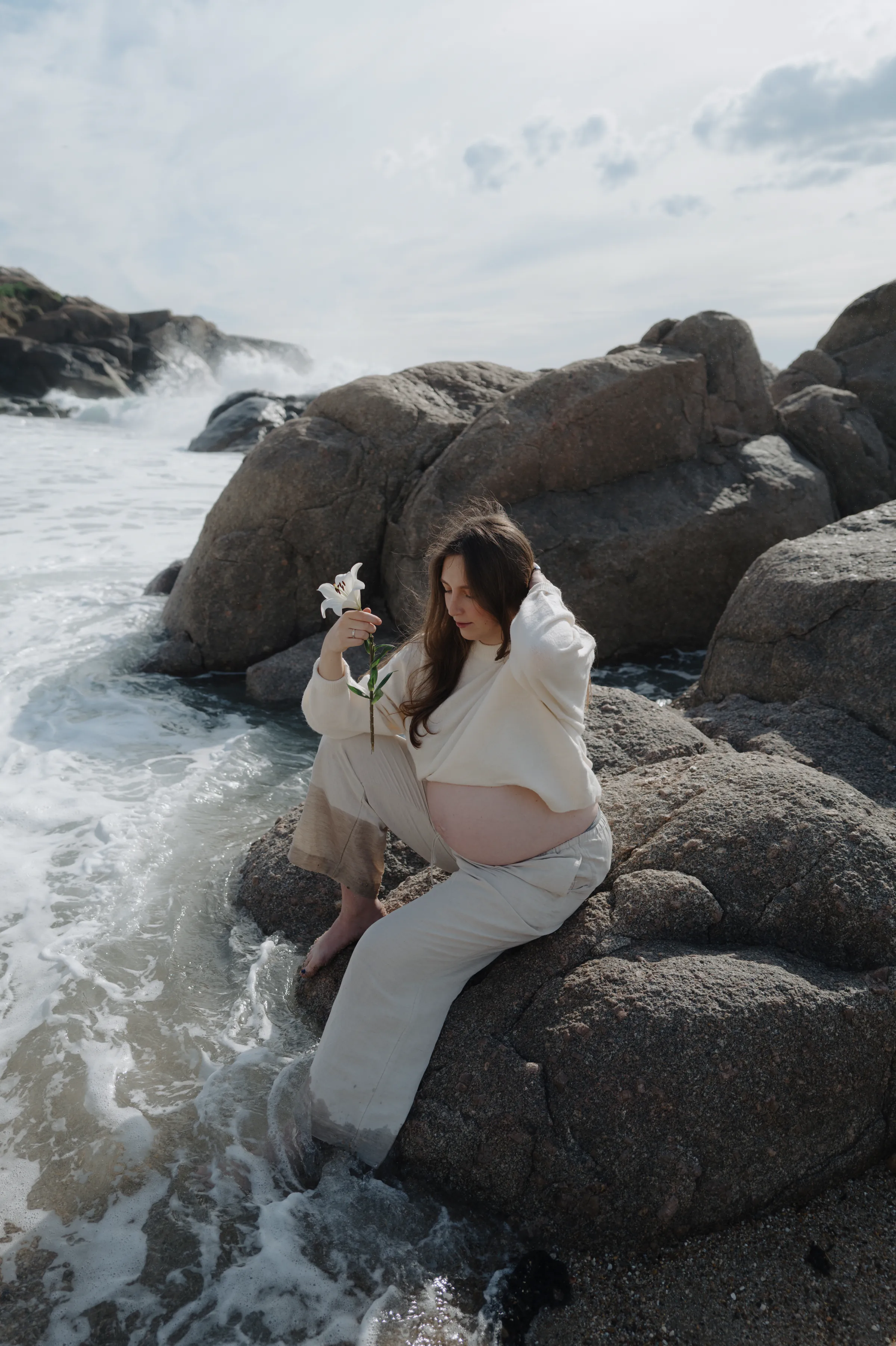 Outdoor seaside portrait of a pregnant person seated on rocks, holding a white flower in soft light.
