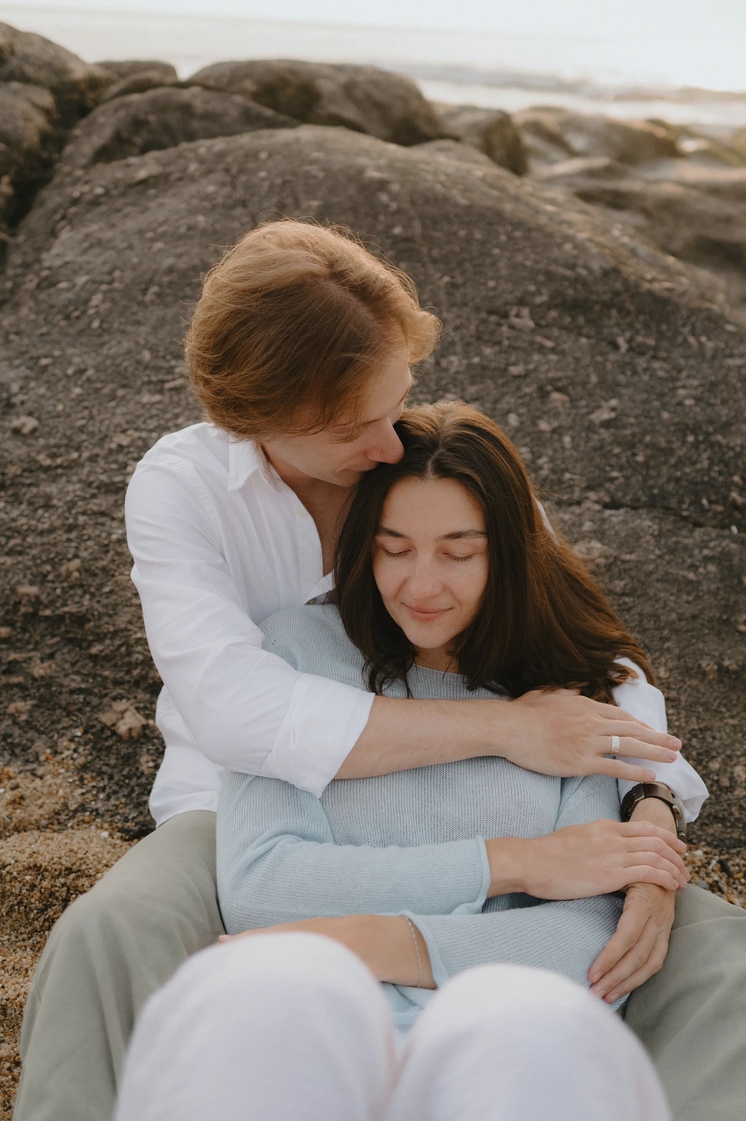Couple seated on seaside rocks in soft light, arms wrapped together for a close portrait.