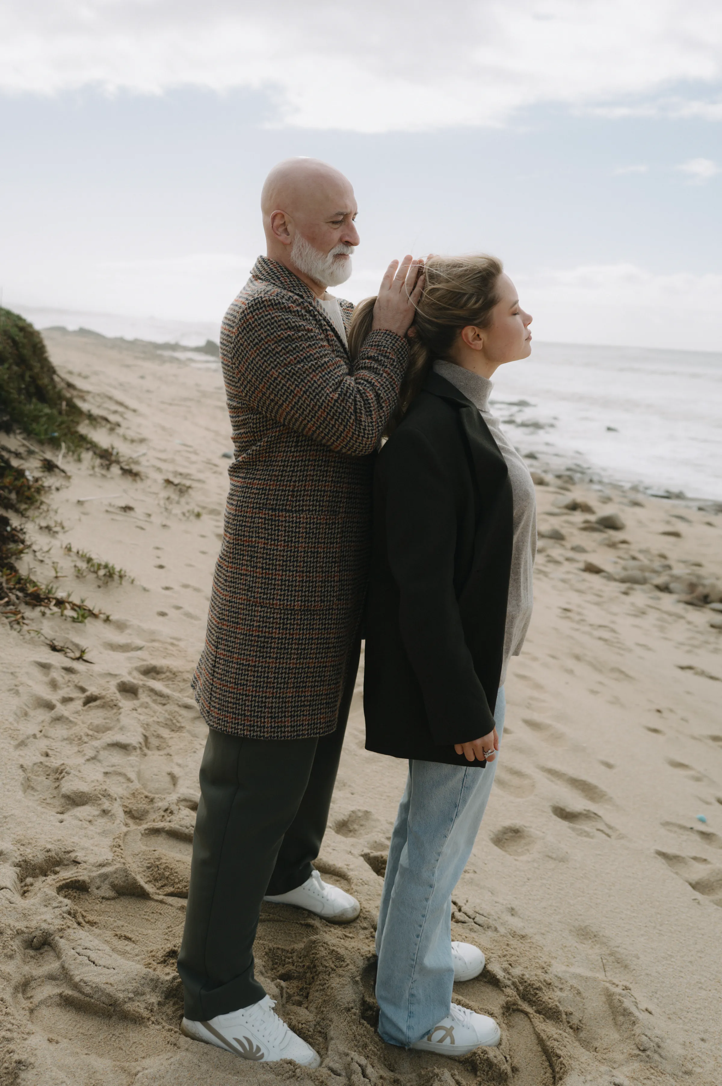 Couple standing close on a sandy seaside outdoor session under soft overcast light.