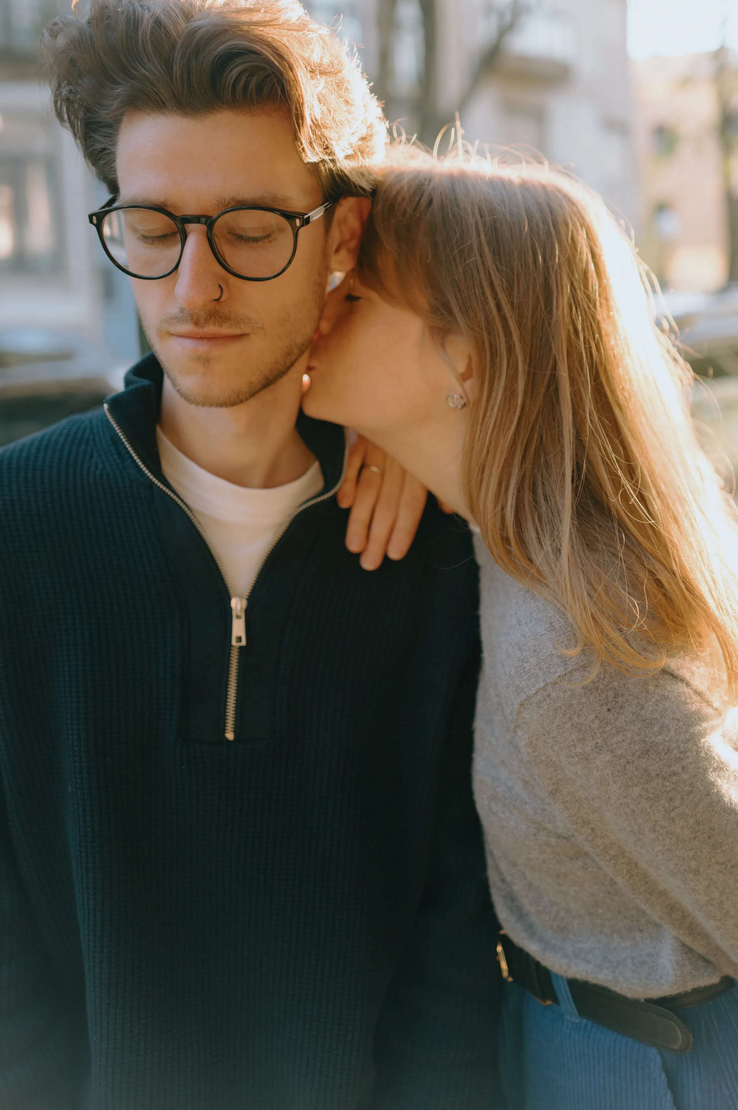 Couple close portrait during a city walk, one person kissing the other's cheek in soft light.
