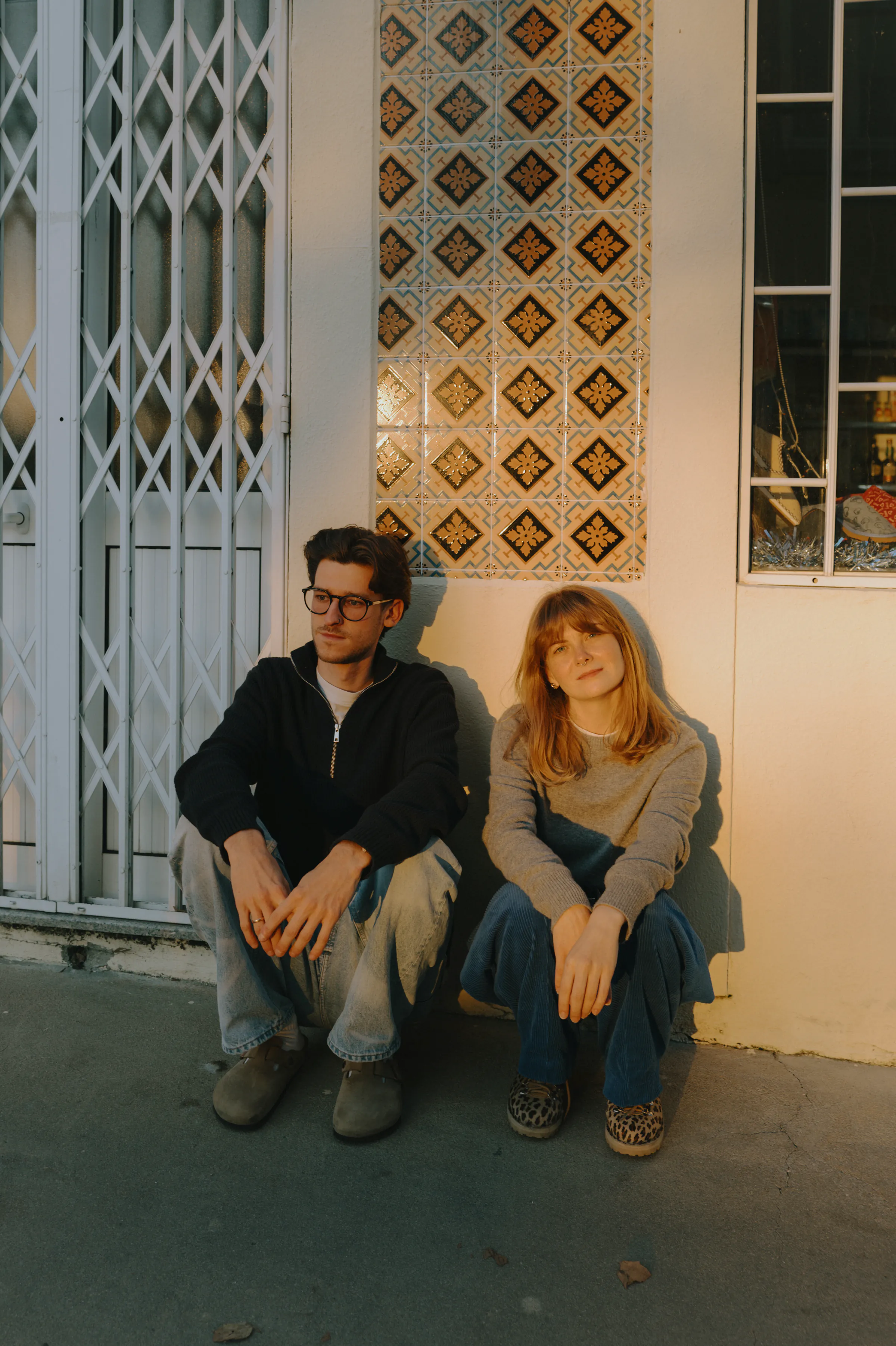 Couple seated on a sidewalk outside a patterned storefront during a warm city walk session.