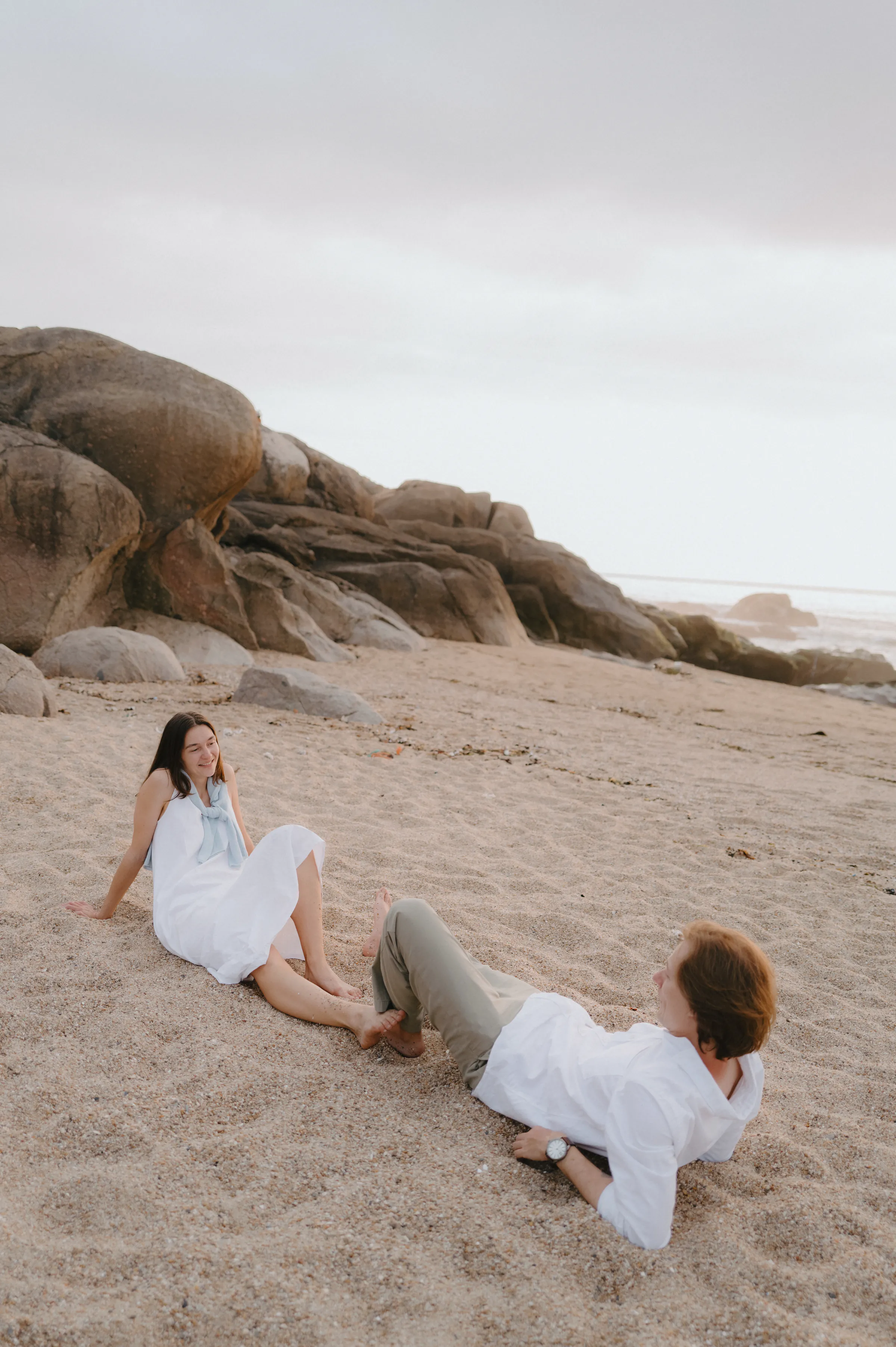 Couple relaxed on a sandy seaside beach beside large rocks in soft overcast light.