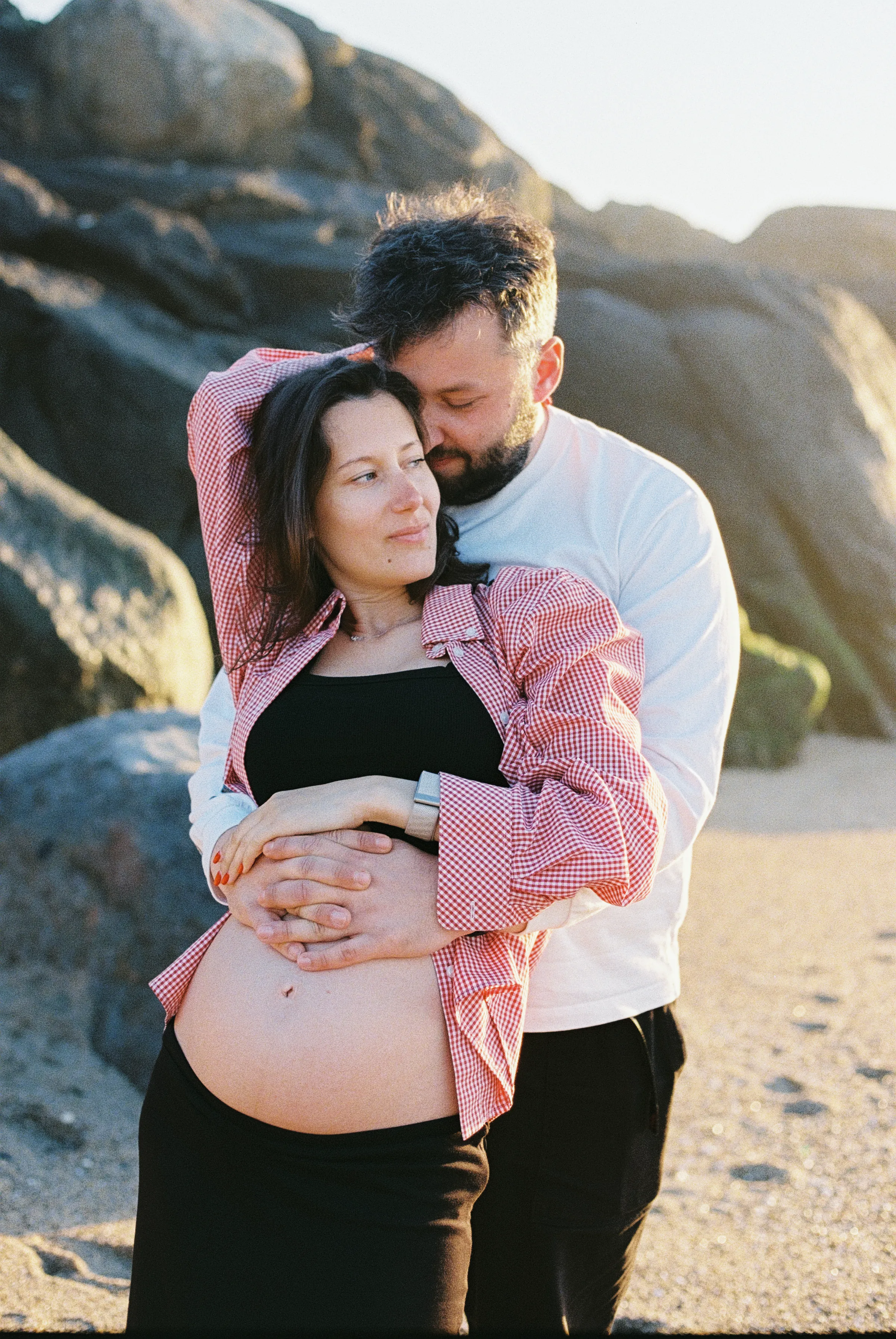 Pregnant couple reclines together on a pebbled beach during a soft light outdoor session.