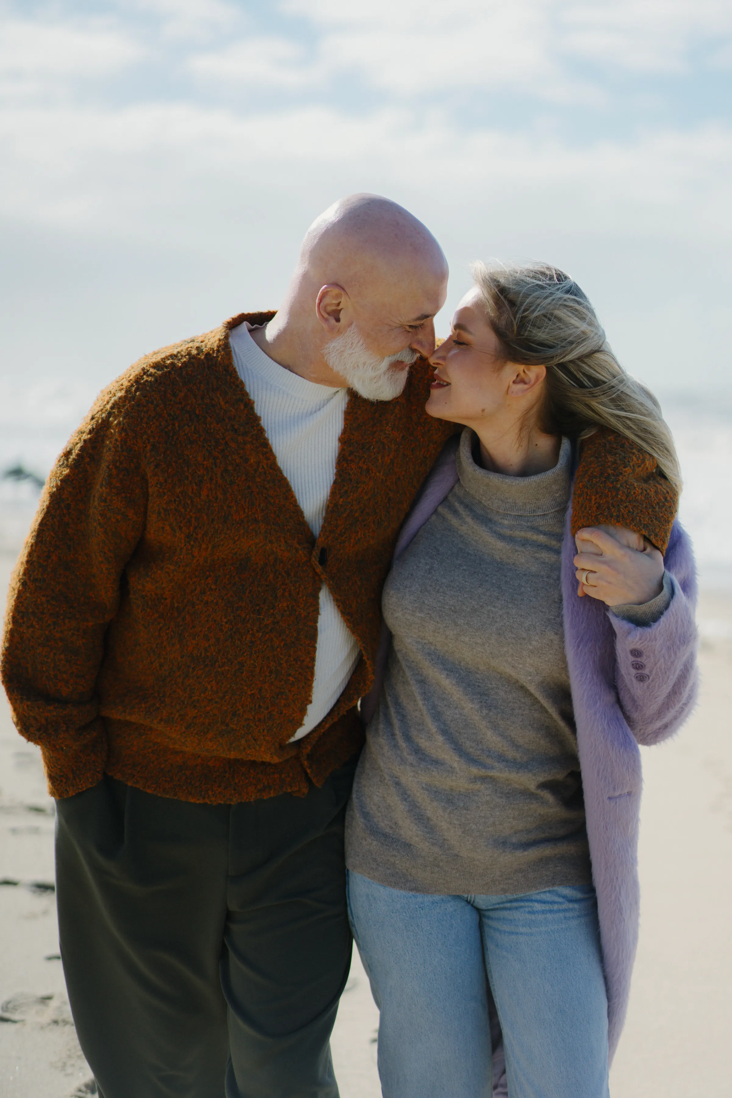Couple walking close together on a seaside session, wrapped in cardigans under soft daylight.