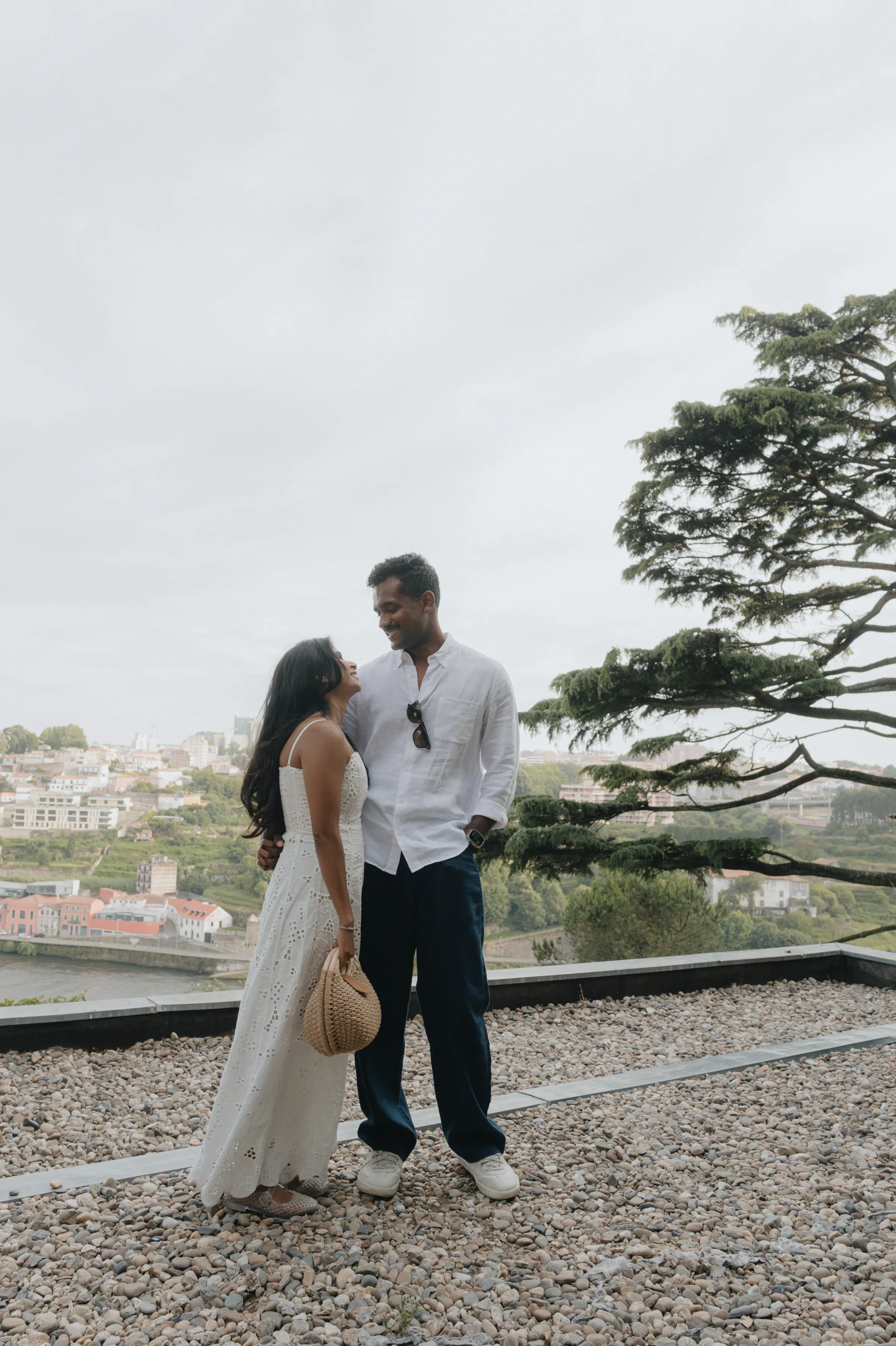 Couple portrait on a gravel terrace overlooking the city, framed by a large tree in soft outdoor light.