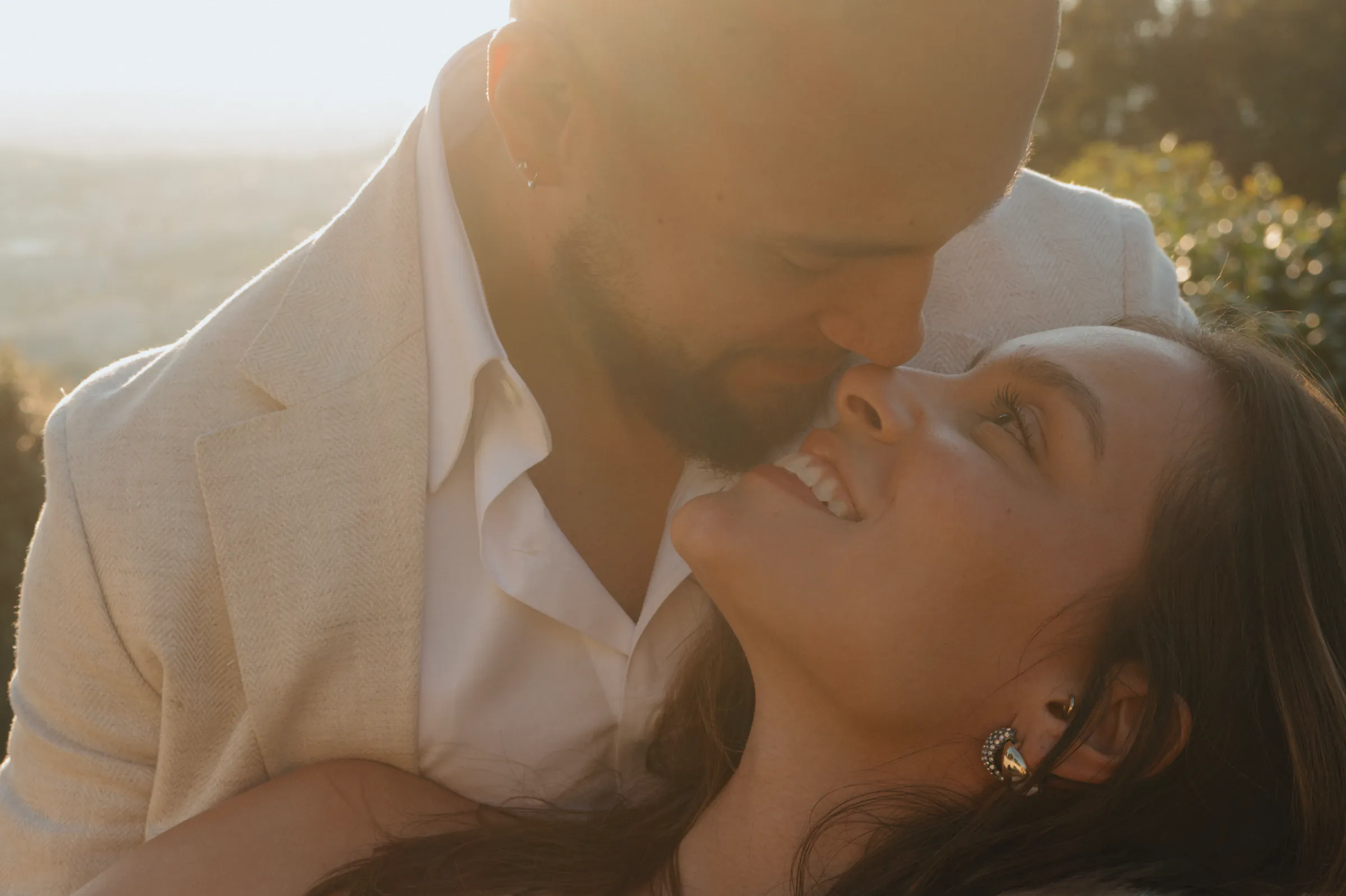 Close portrait of a couple leaning together outdoors in soft light, with greenery blurred behind them.