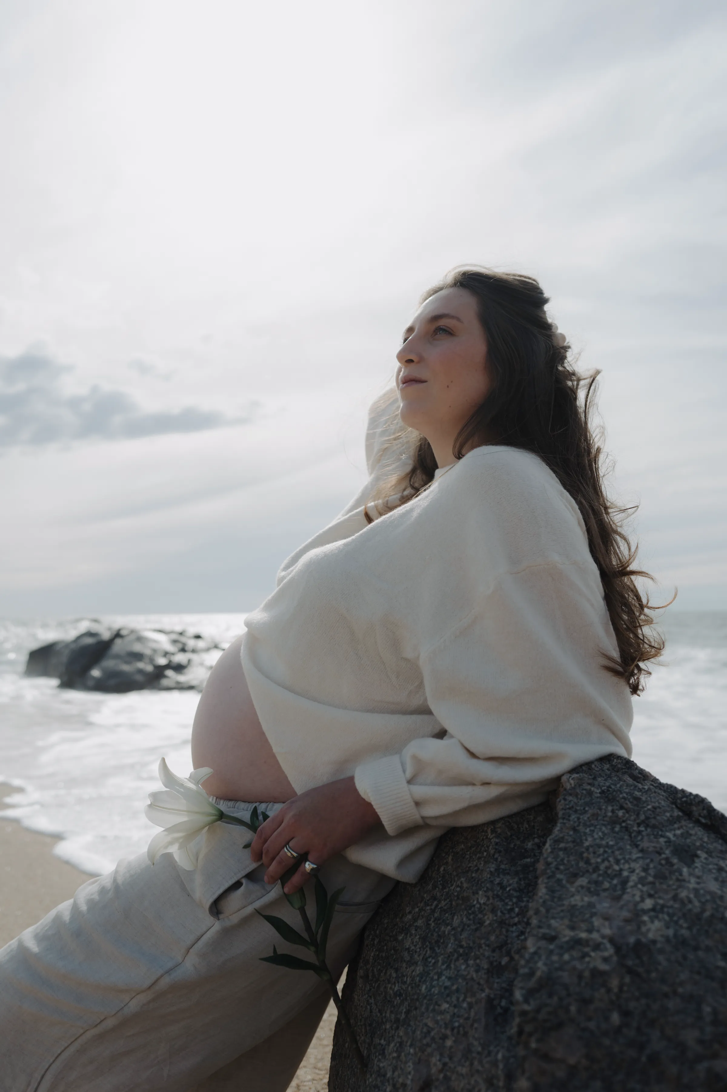 Seaside portrait of a person in white leaning against rocks, holding flowers in soft light.