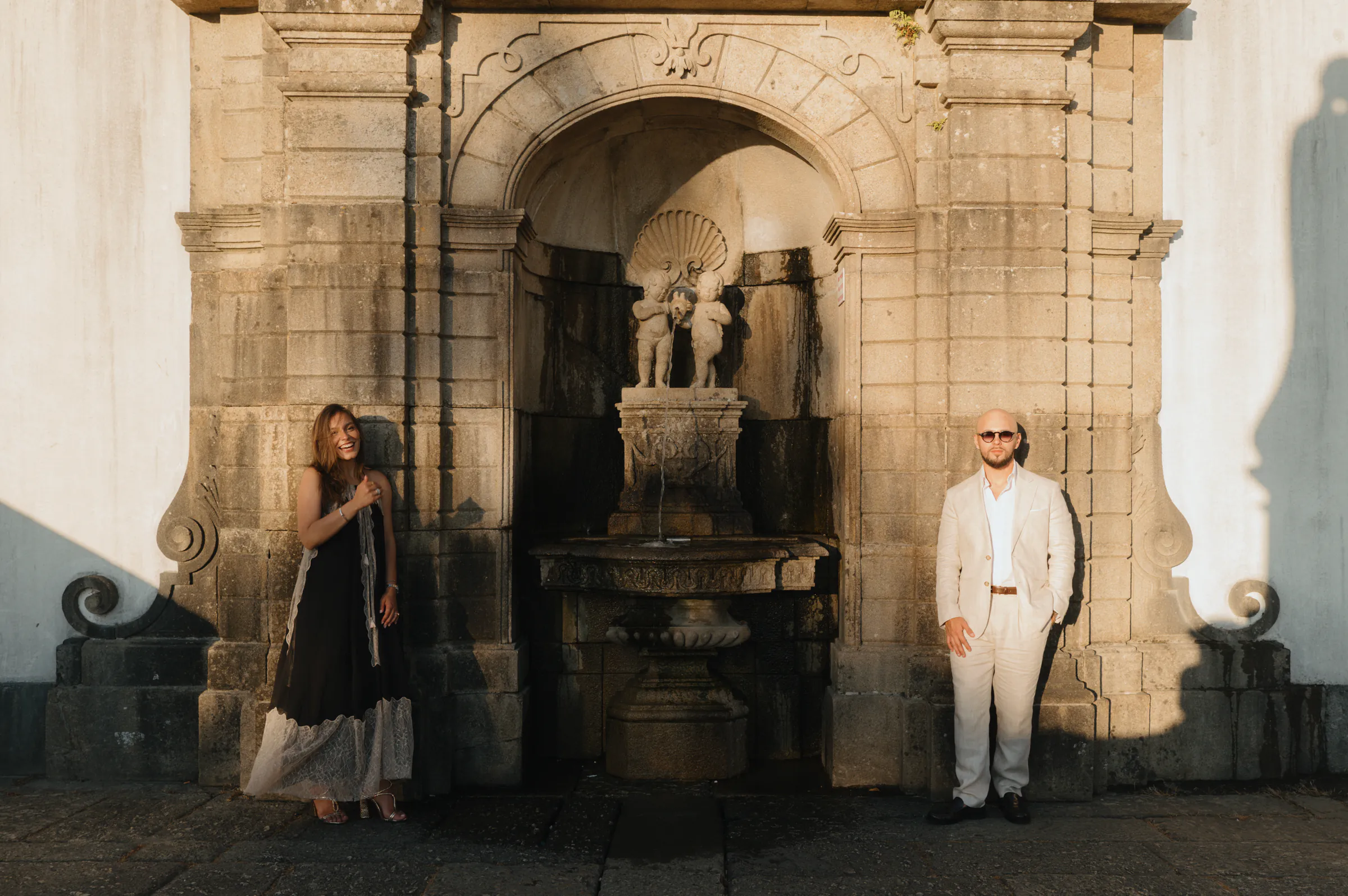 Outdoor portrait of two adults posed by an ornate stone archway and fountain in soft light.