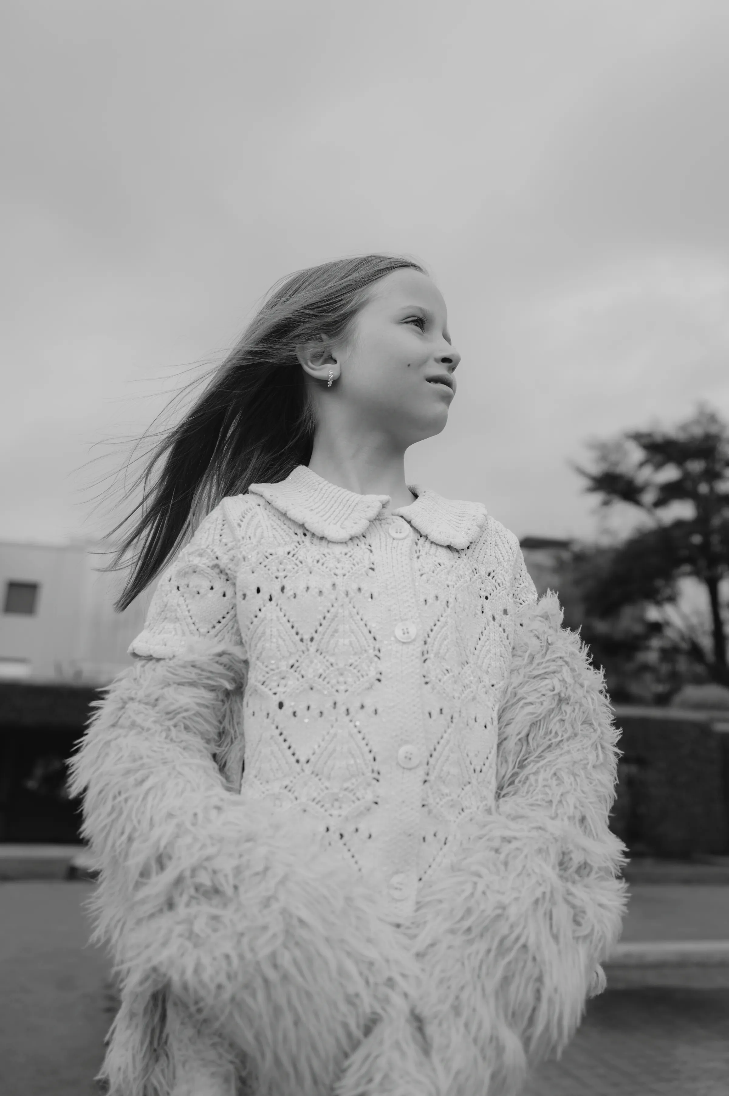 Black-and-white child portrait outdoors, wearing a textured sweater and looking aside under a cloudy sky.