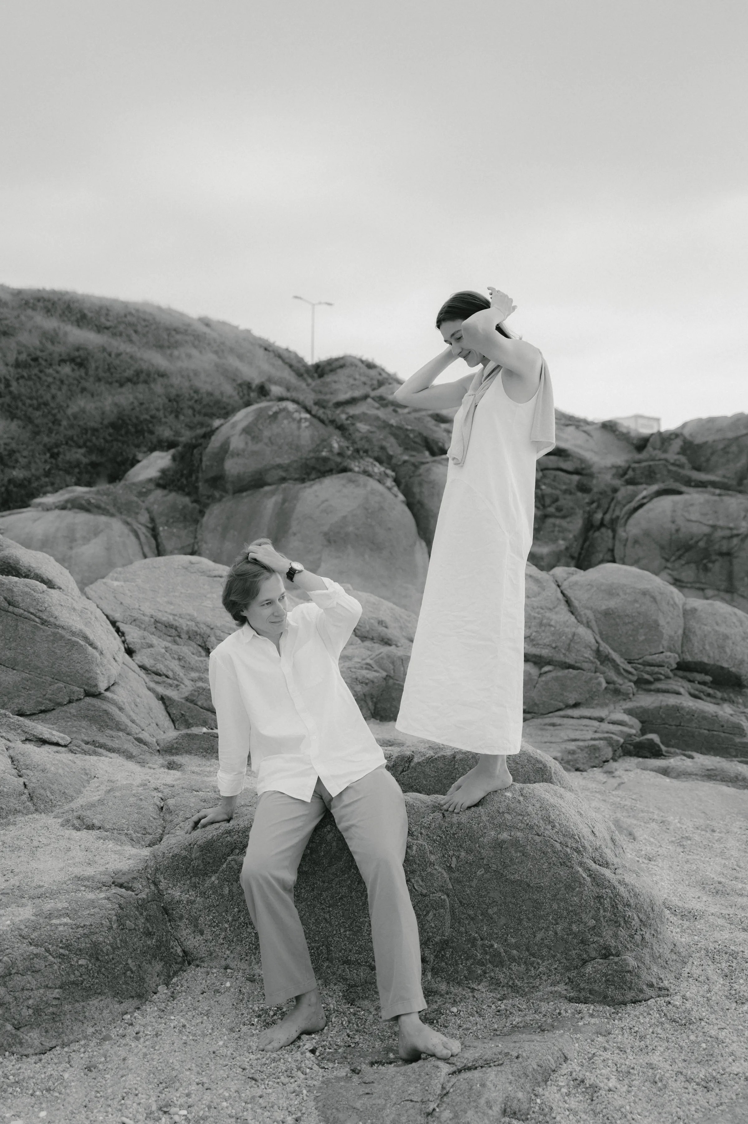 Couple in white outfits during a black-and-white outdoor session on rocky coastal terrain.