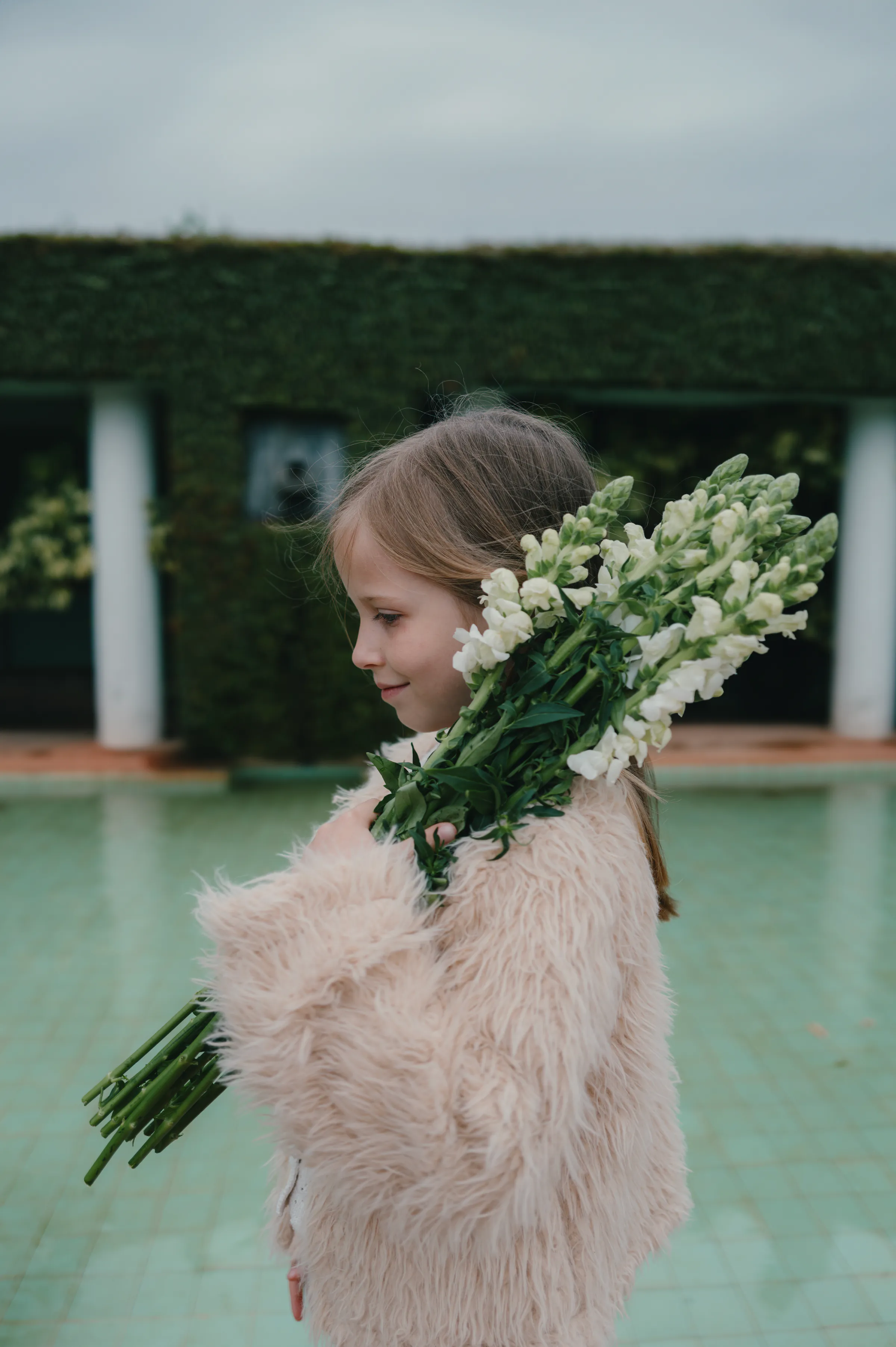 Child portrait holding white flowers during an outdoor session by a pool in soft overcast light.