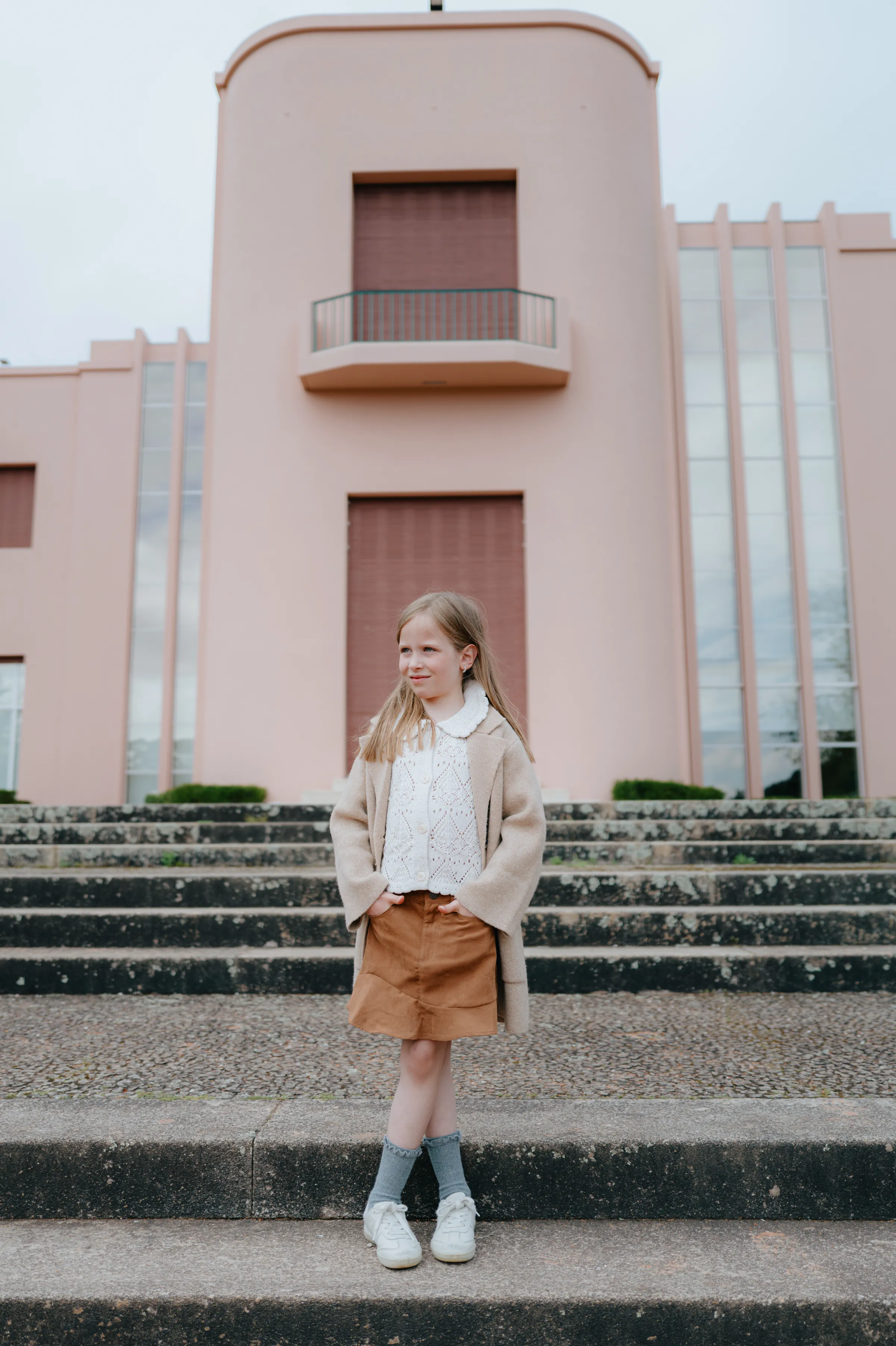 Child portrait on outdoor steps in soft light, framed by a pale pink building and tall windows.