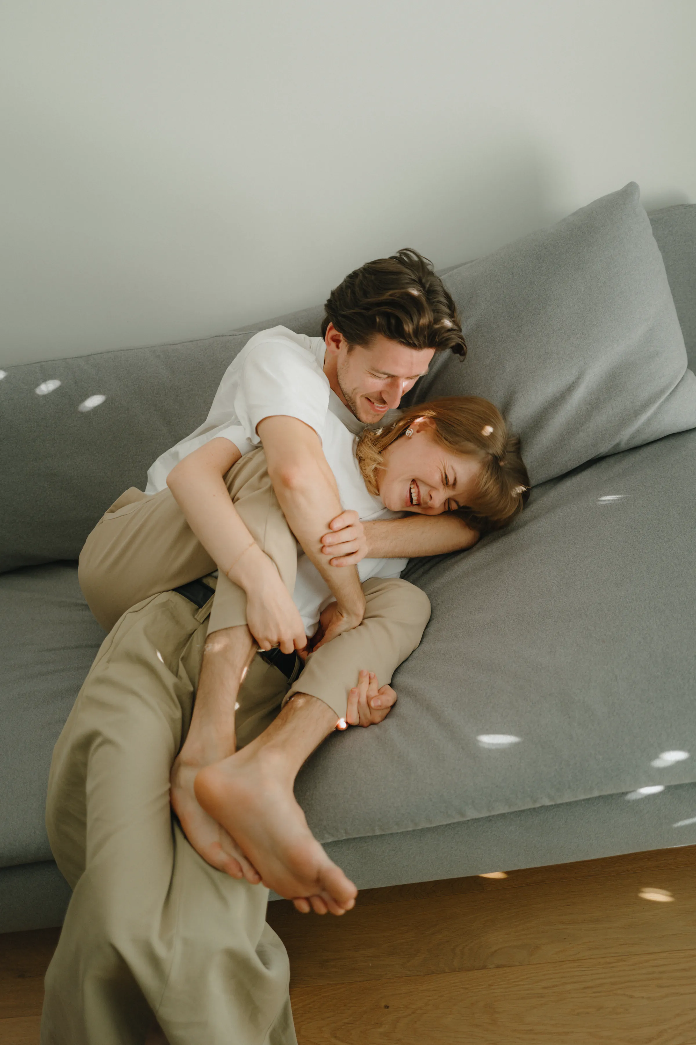 Couple cuddling on a gray sofa during a relaxed home session in soft window light.