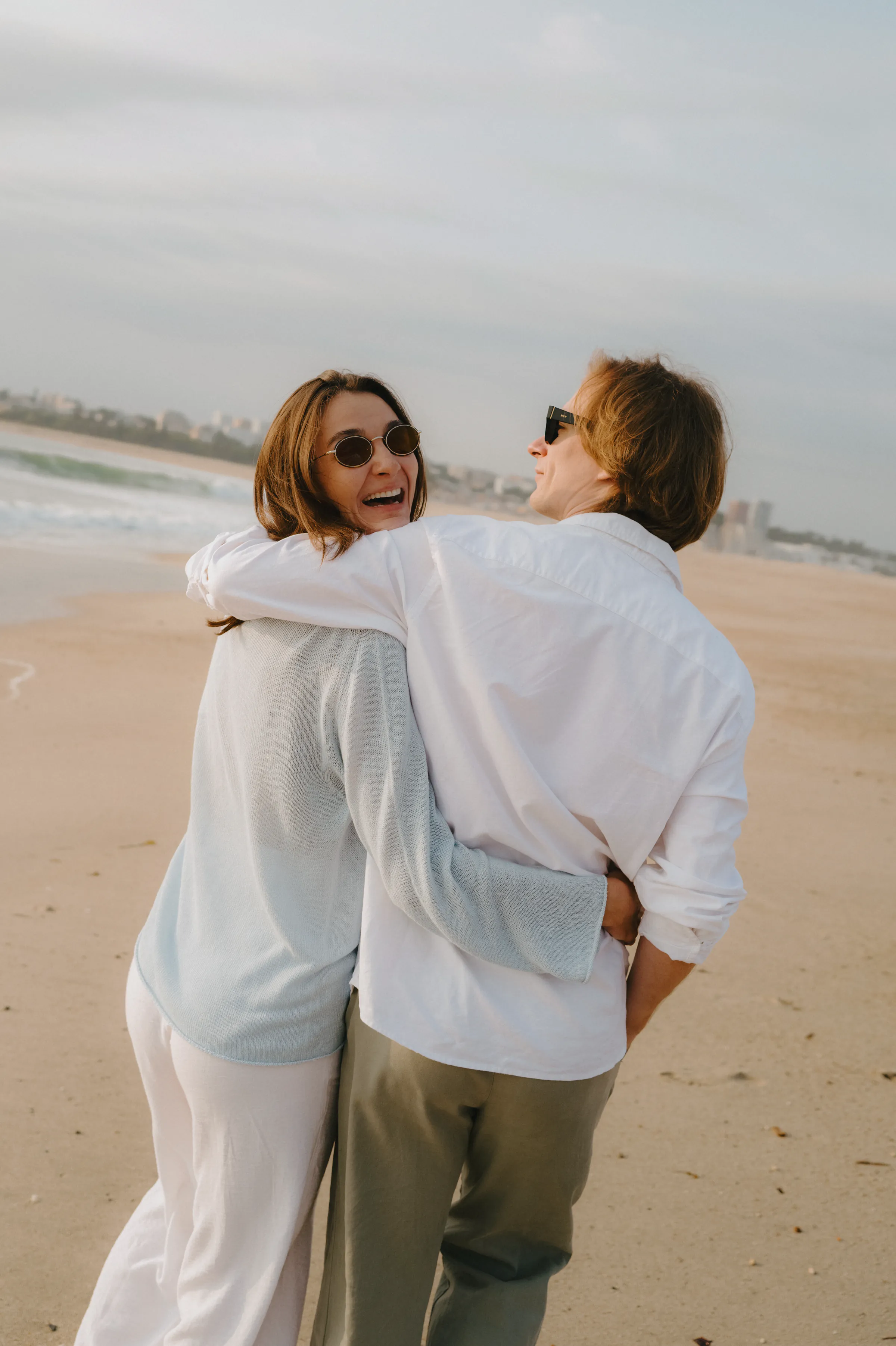 Couple wearing sunglasses walk arm in arm along a sandy beach during a soft seaside session.