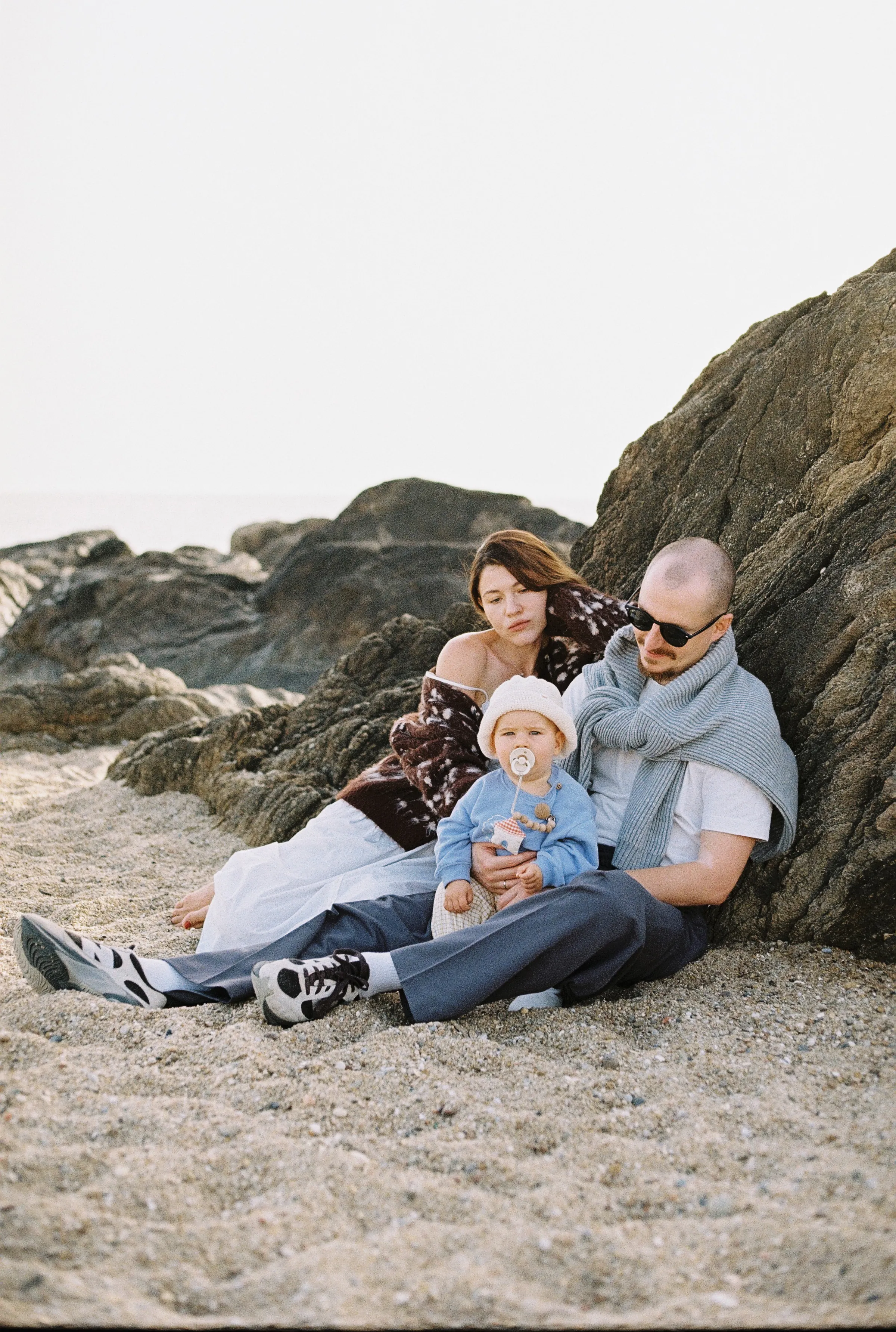 Family portrait with baby seated on a sandy seaside beach beside large rocks in soft light