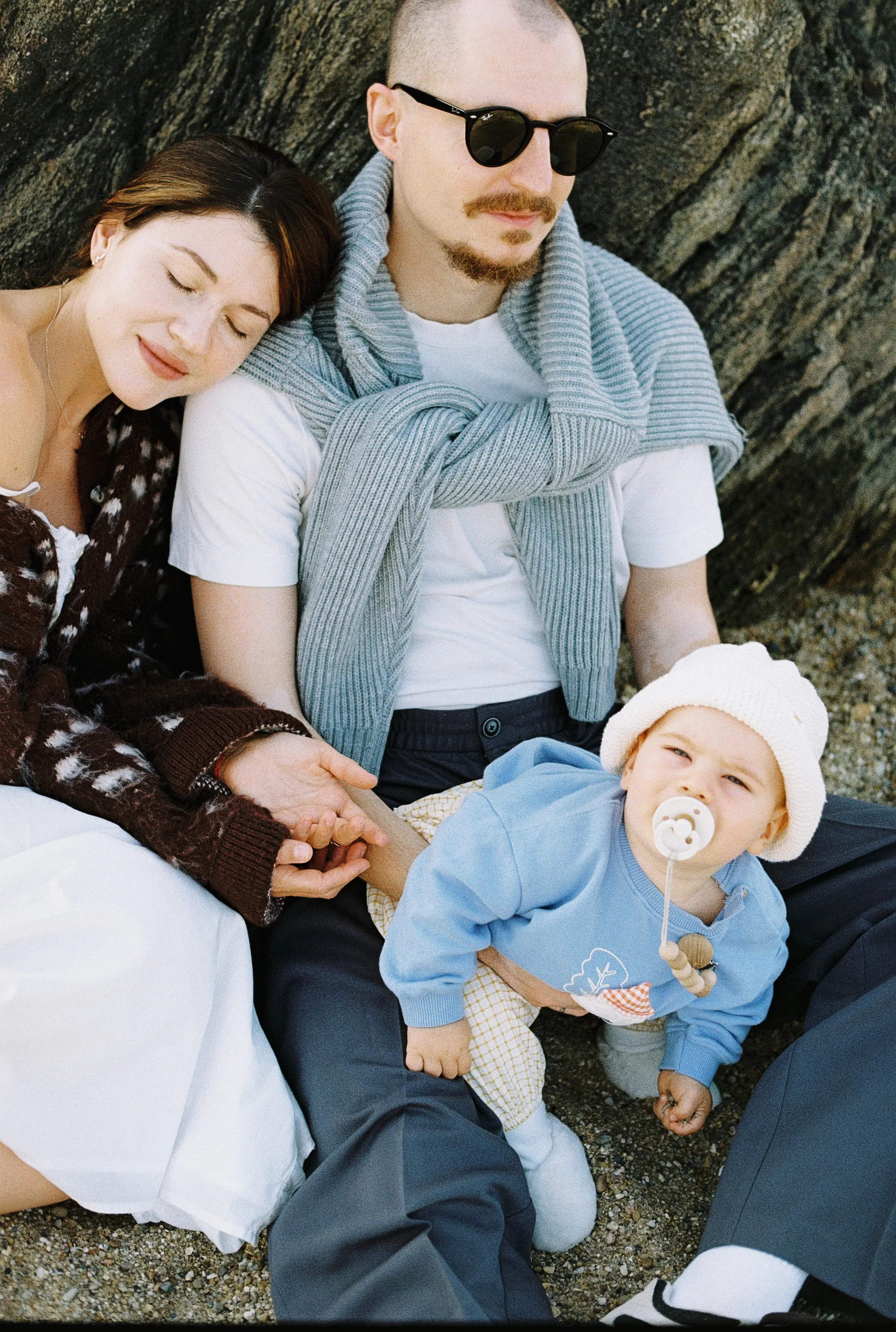 Family outdoor session with a couple seated close together beside a baby in soft light.