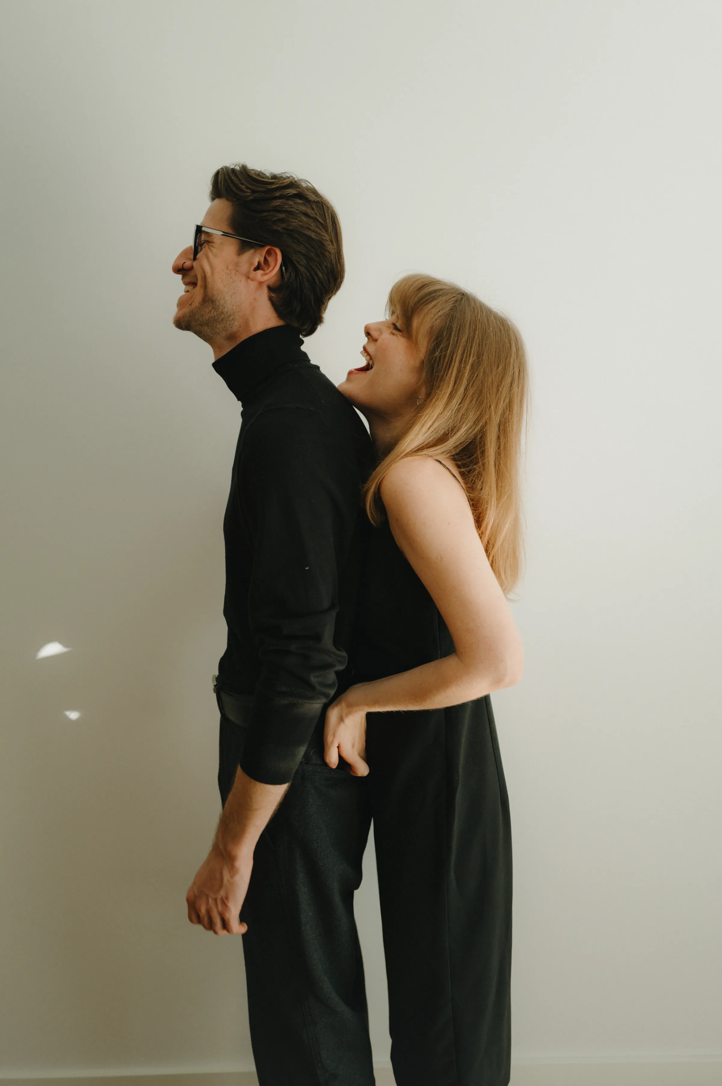 Couple portrait indoors, standing close in black outfits against a plain wall.
