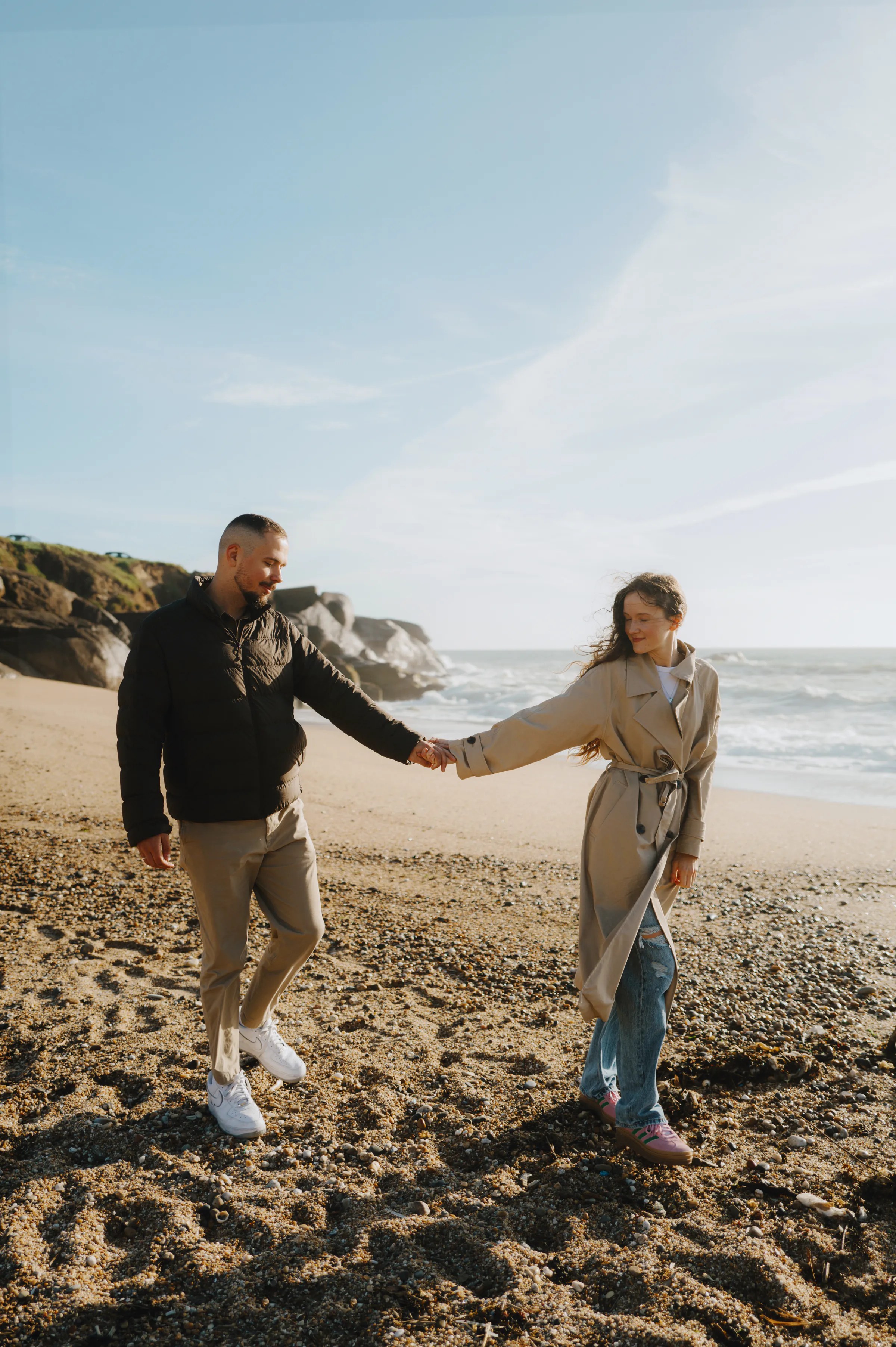 Couple holding hands during a seaside outdoor session on a pebbly beach with cliffs and soft light.