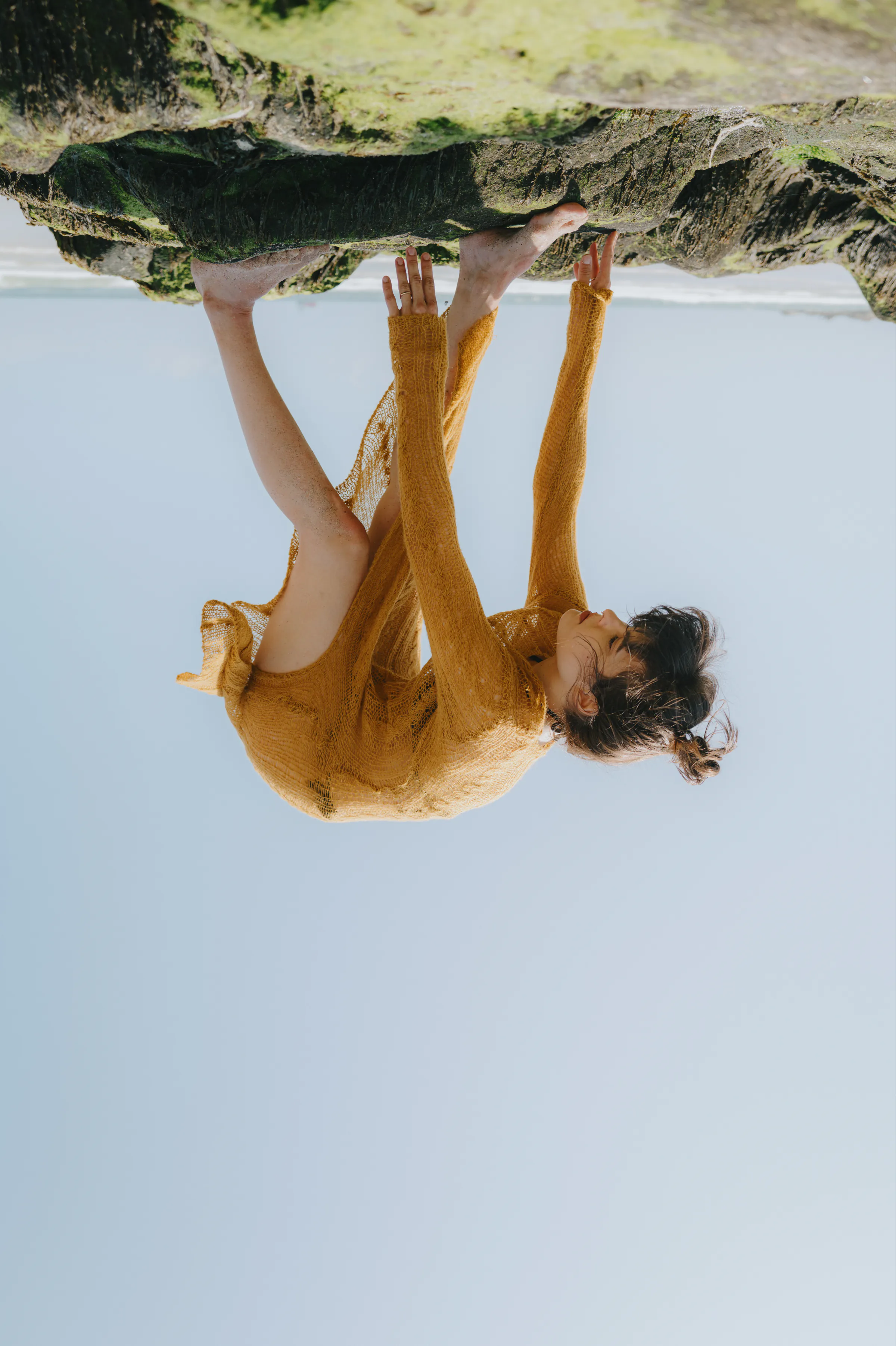 Outdoor portrait of a person in a mustard dress posing on rocky seaside cliffs under clear sky.