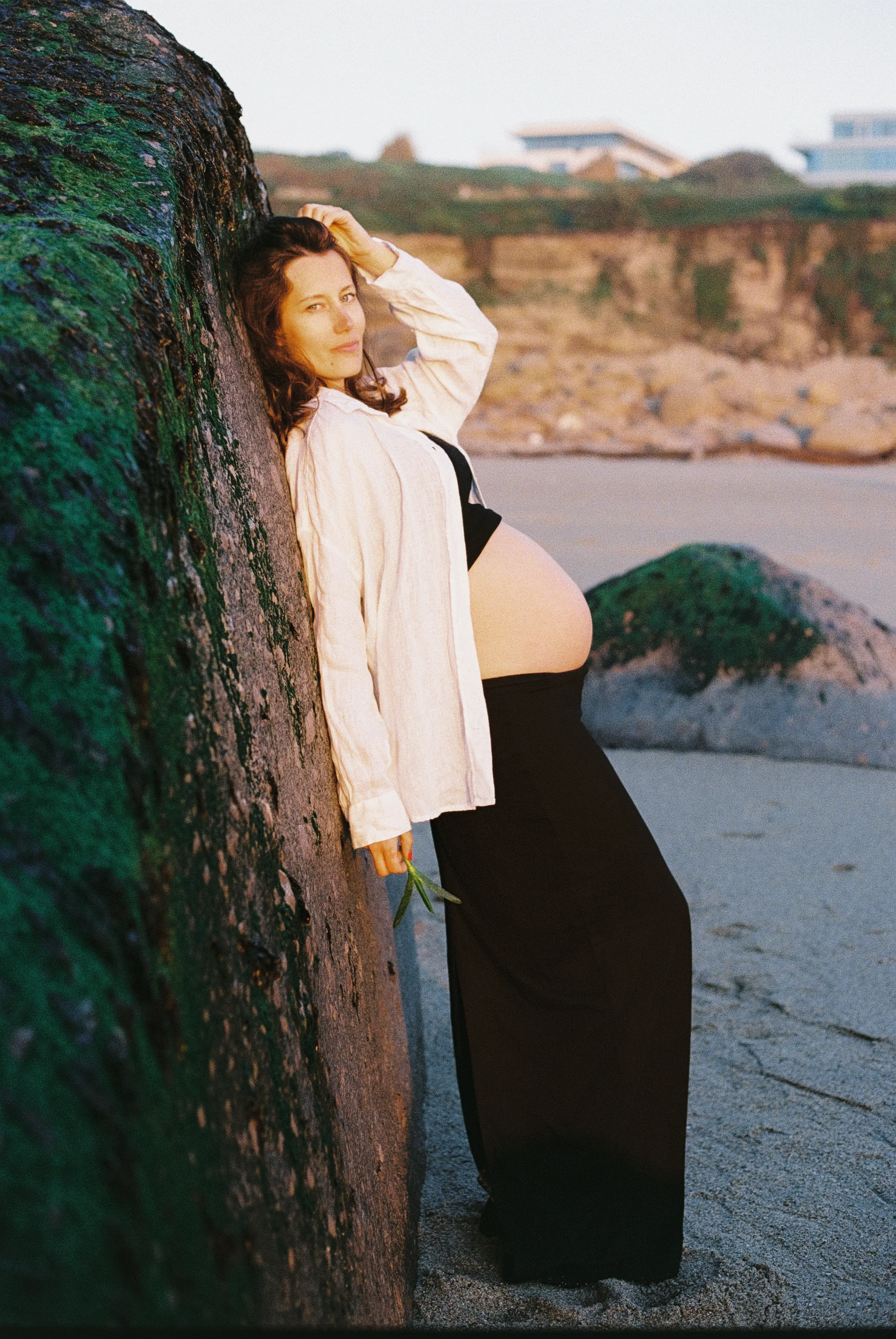 Pregnant person leaning against a large seaside rock during an outdoor portrait session in soft light.