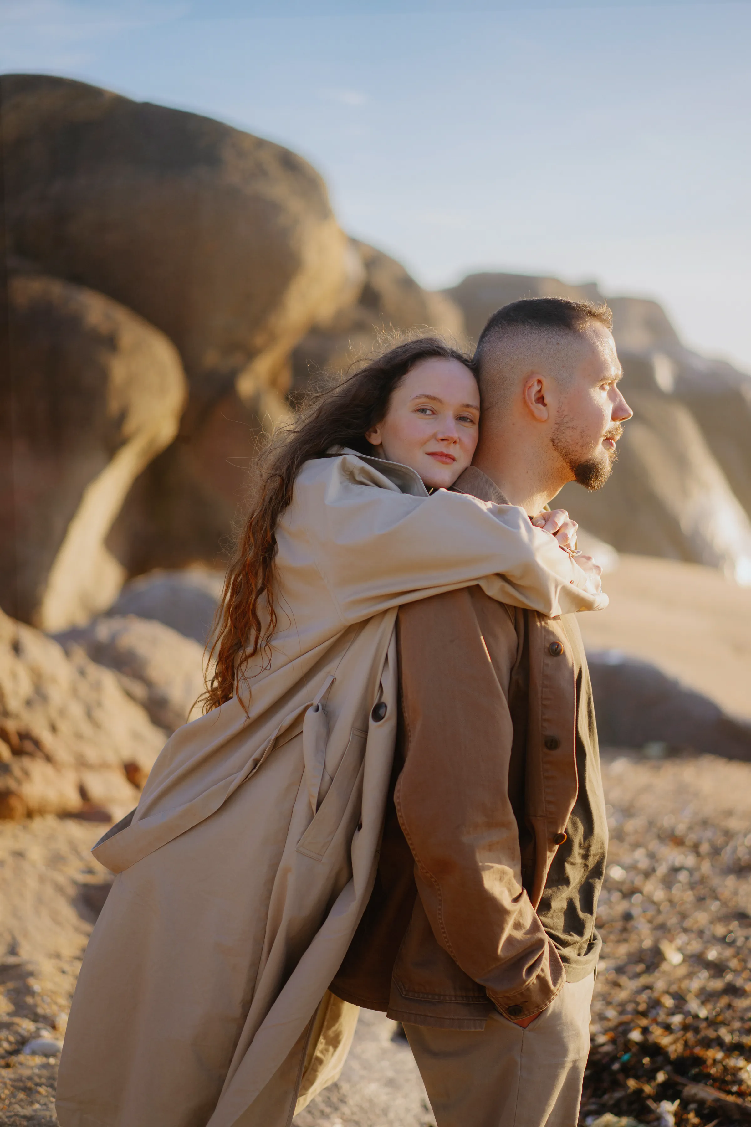 Couple portrait during a seaside outdoor session, standing close among coastal rocks in soft light.