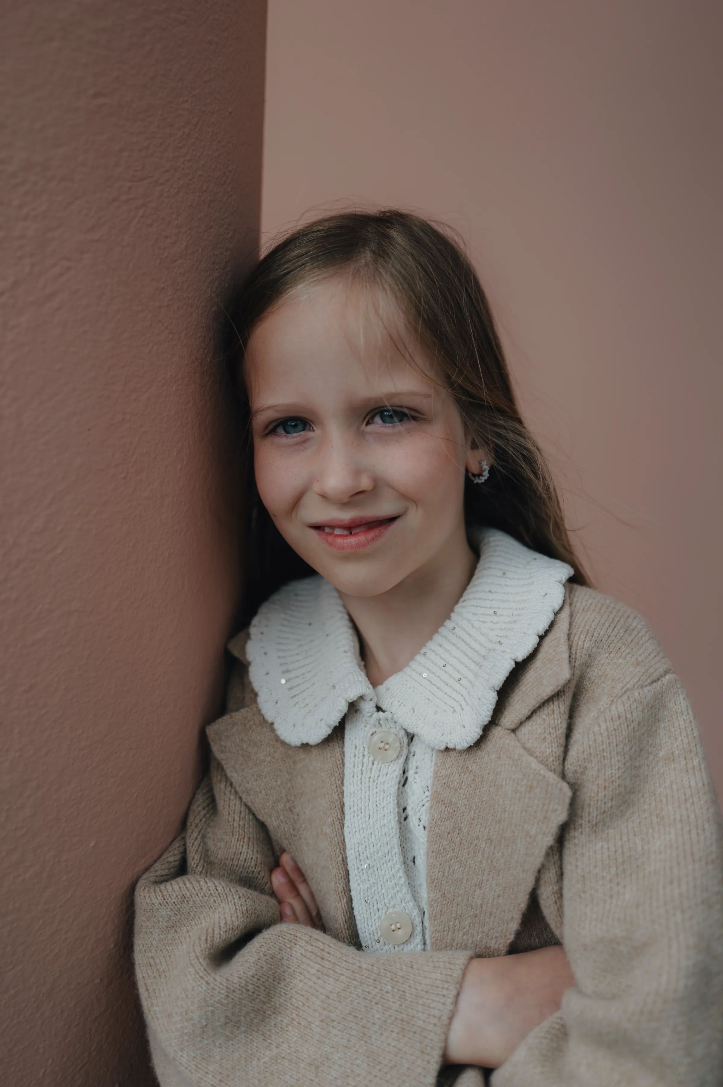 Child portrait by a muted wall, wearing a beige coat and white knit collar in soft light.