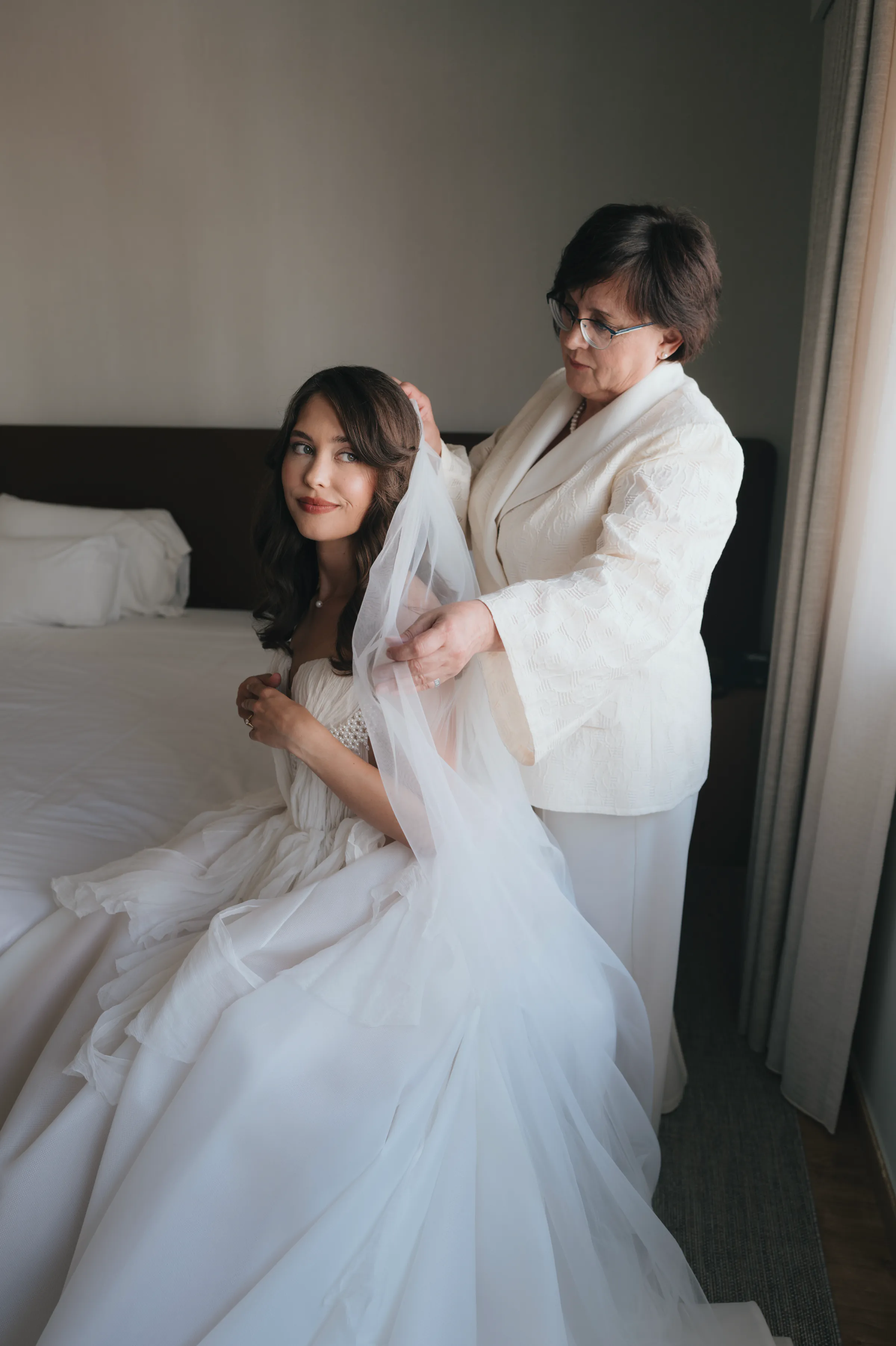 Indoor bridal portrait as an older adult adjusts a seated woman's veil beside a bed in soft window light.