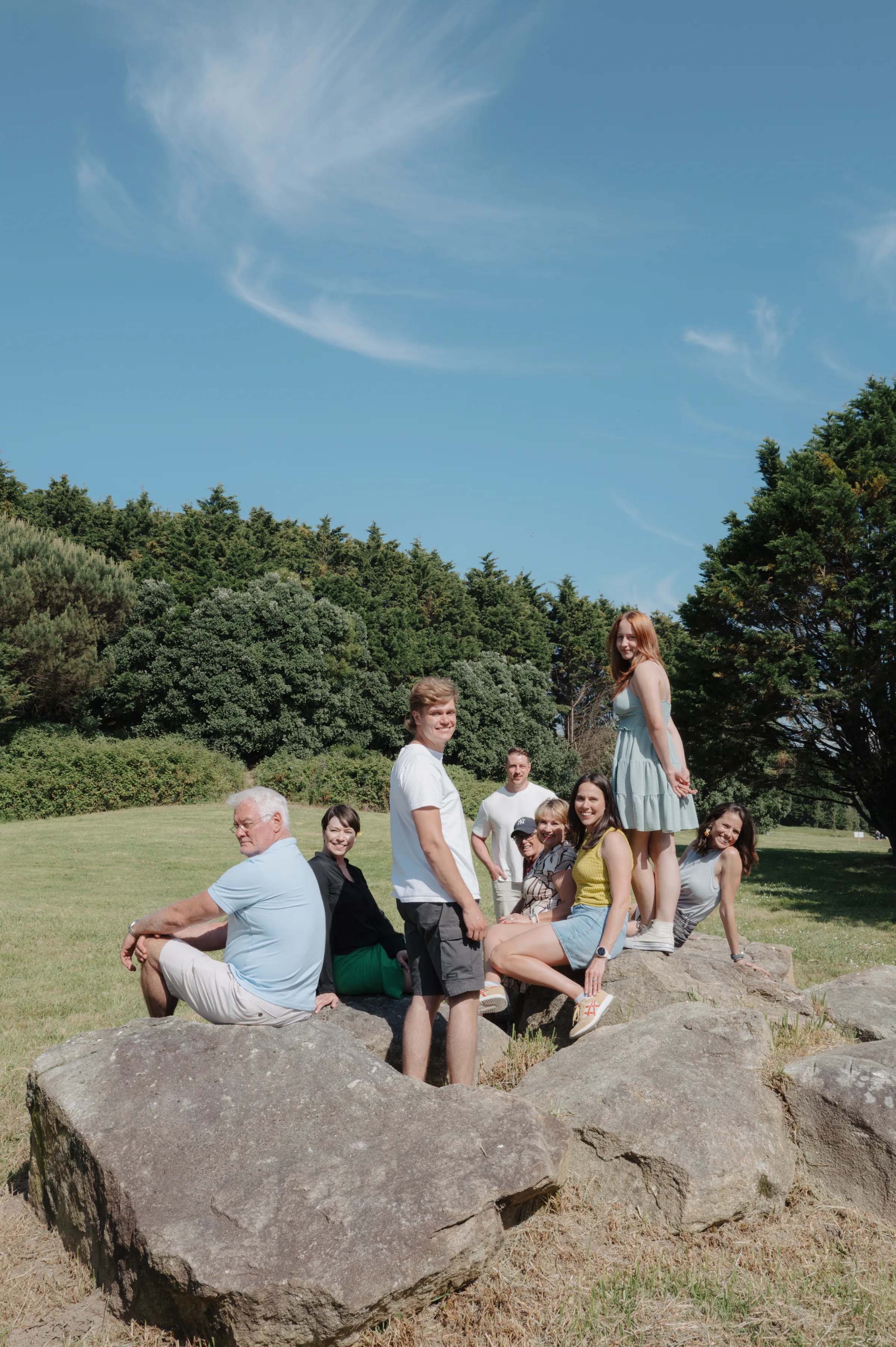 Family outdoor session on large rocks in a grassy park, with trees and open sky behind them.
