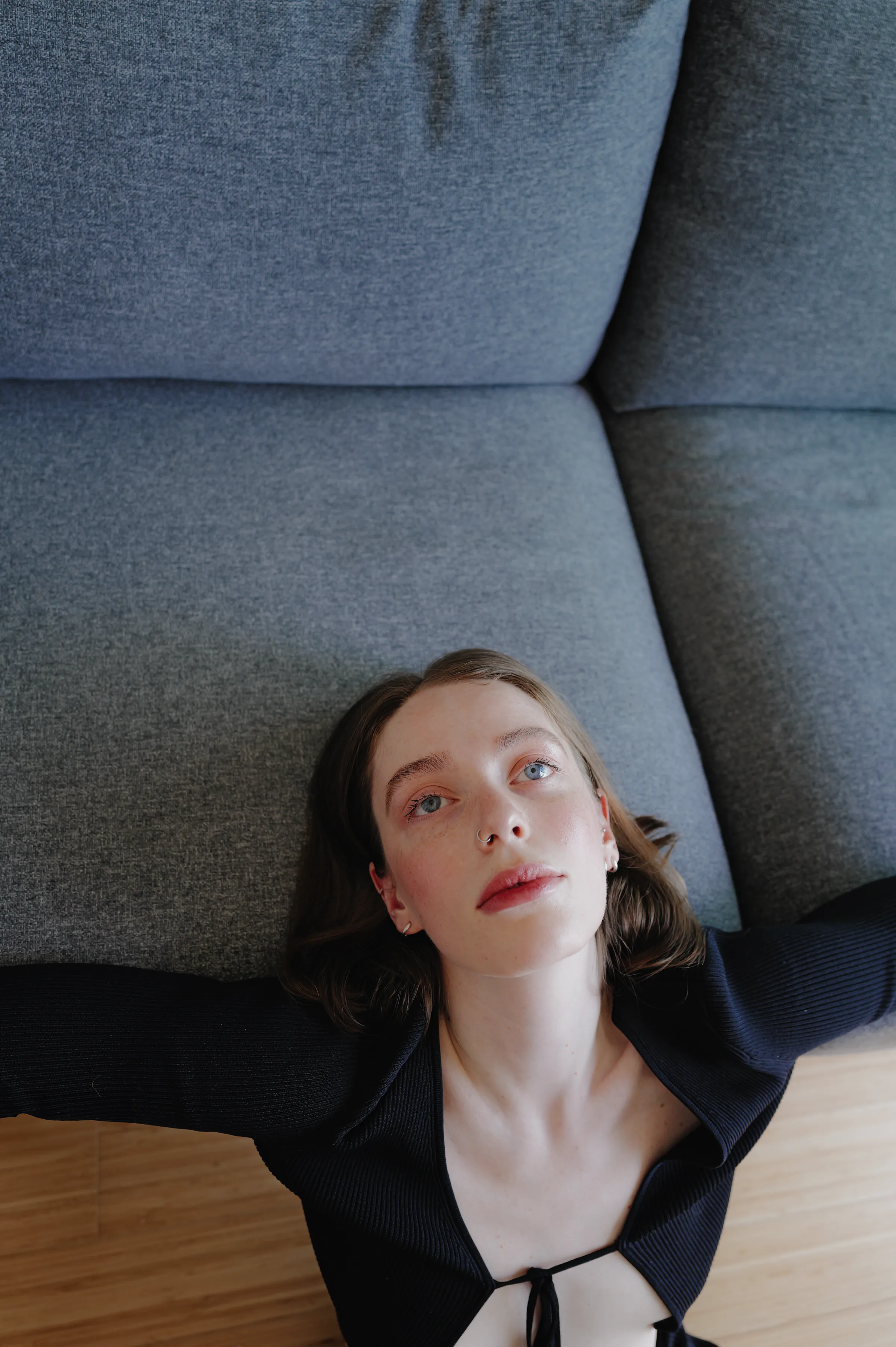 Close portrait of a woman reclining beneath gray sofa cushions in soft indoor light.