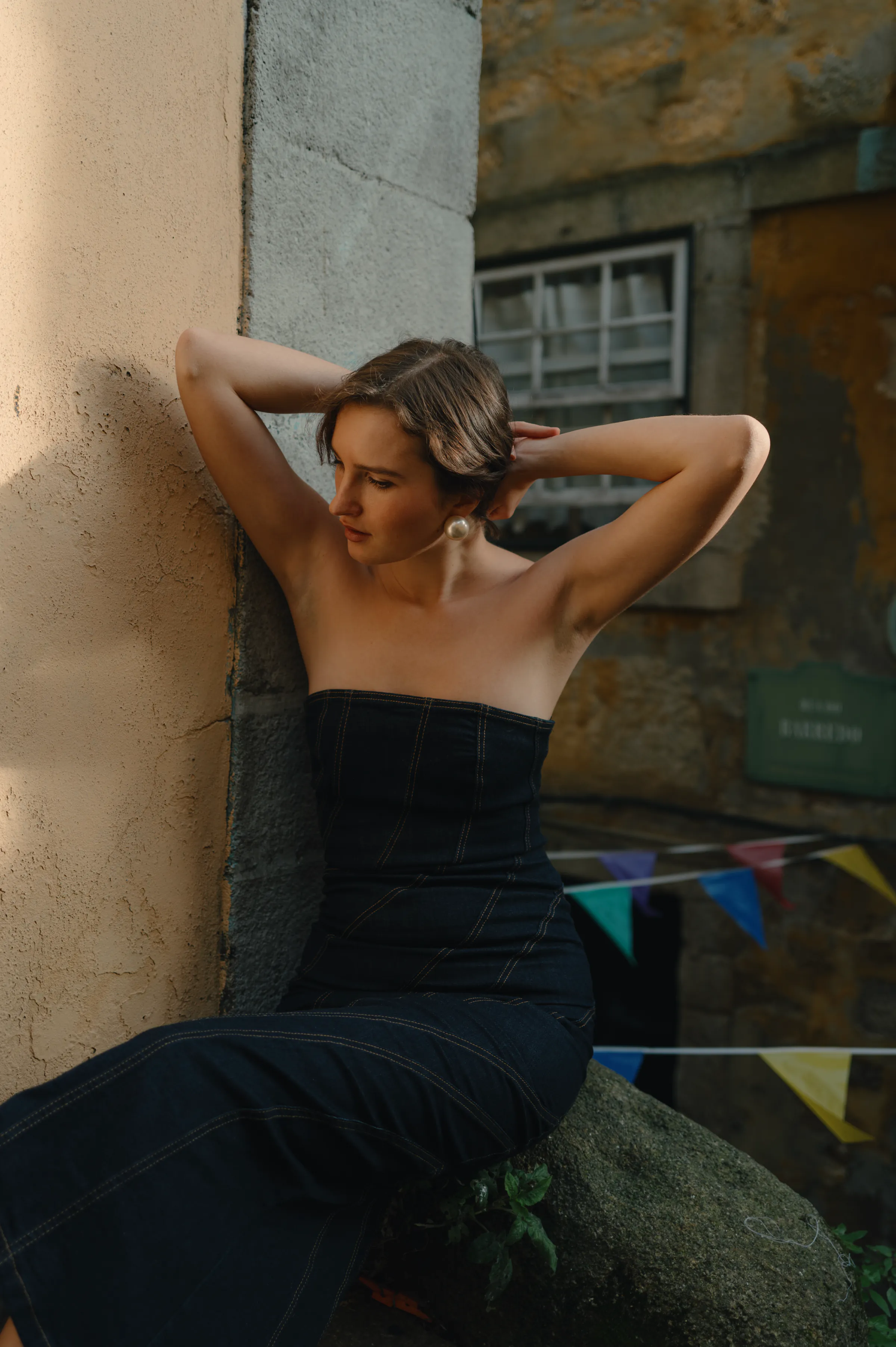 Outdoor portrait of a person in a dark strapless dress leaning by a textured stone wall in soft light.