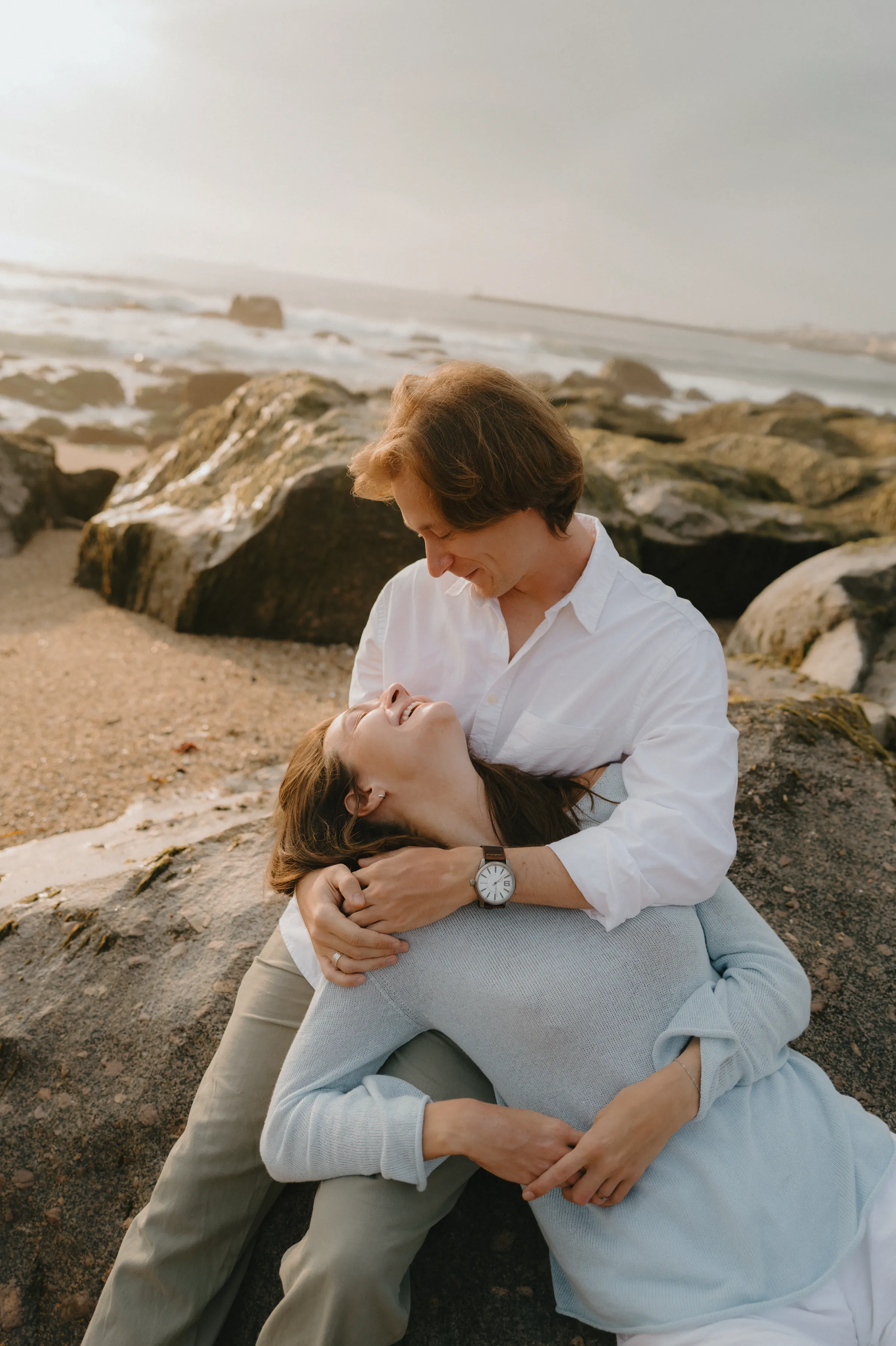 Couple portrait on seaside rocks, sitting close together in soft light beside the waves.