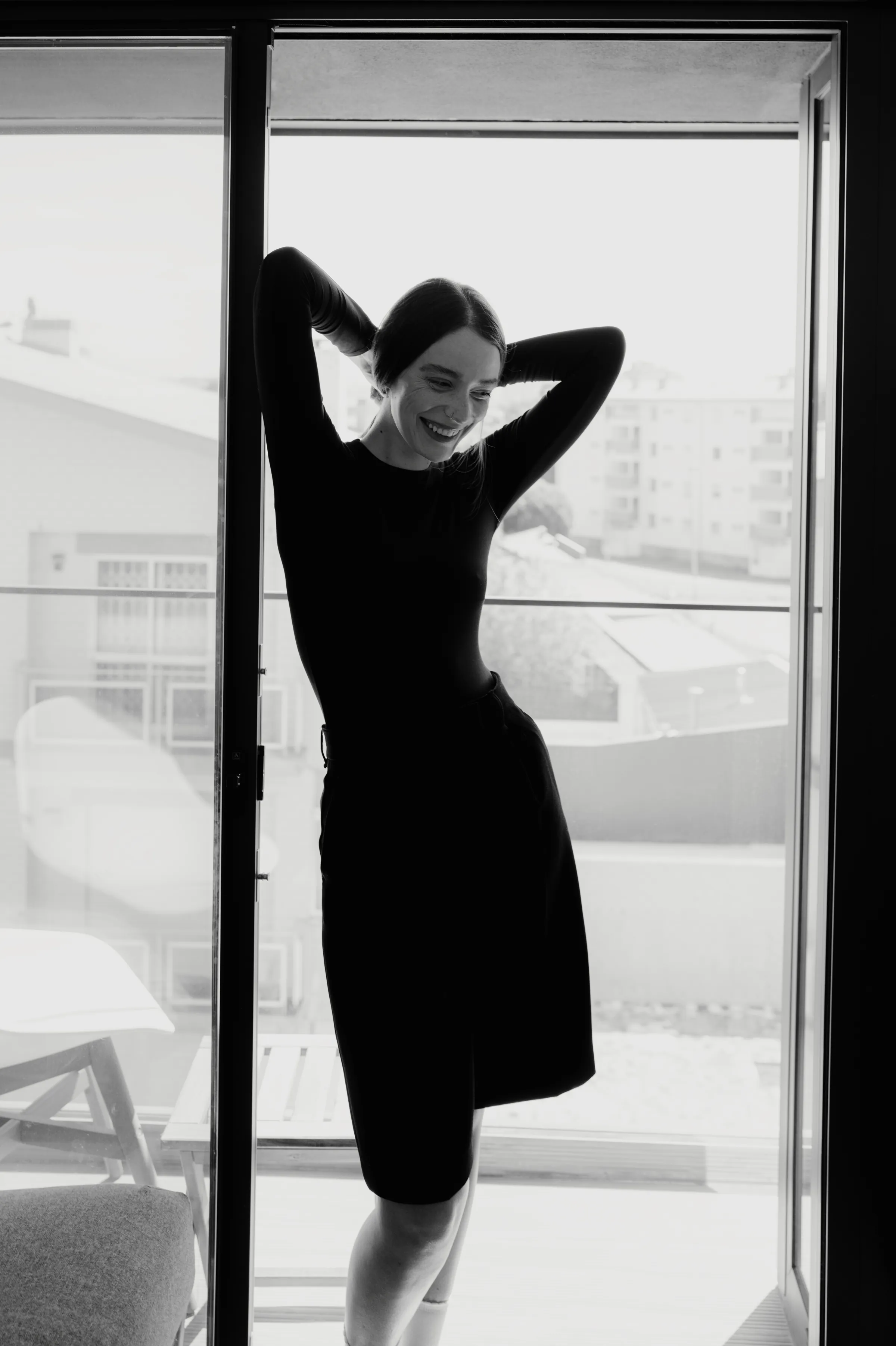 Black-and-white portrait by a balcony door, with soft window light and city buildings beyond.
